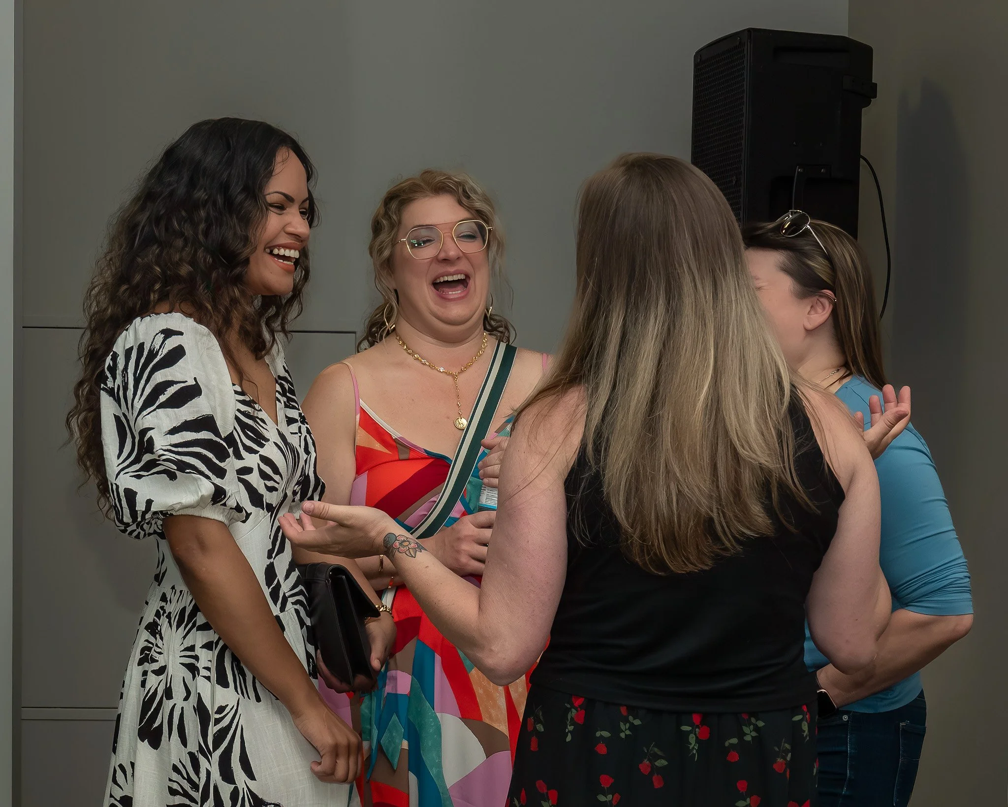 A group of women engaged in conversation, smiling and laughing, with a plain wall and a speaker in the background.