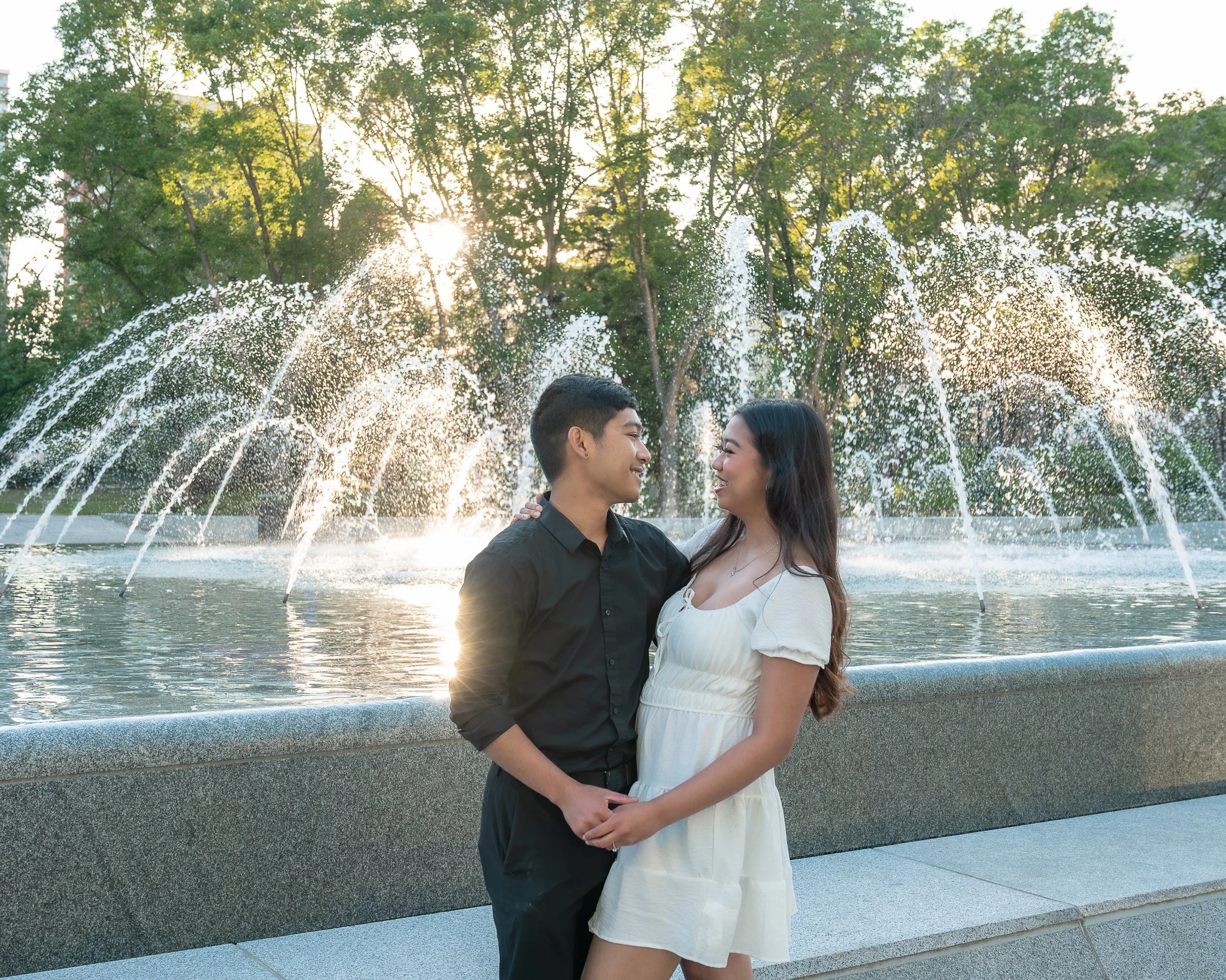 A young couple stands close together near a fountain in a park during sunset, smiling and gazing into each other's eyes, with trees and sunlight in the background.