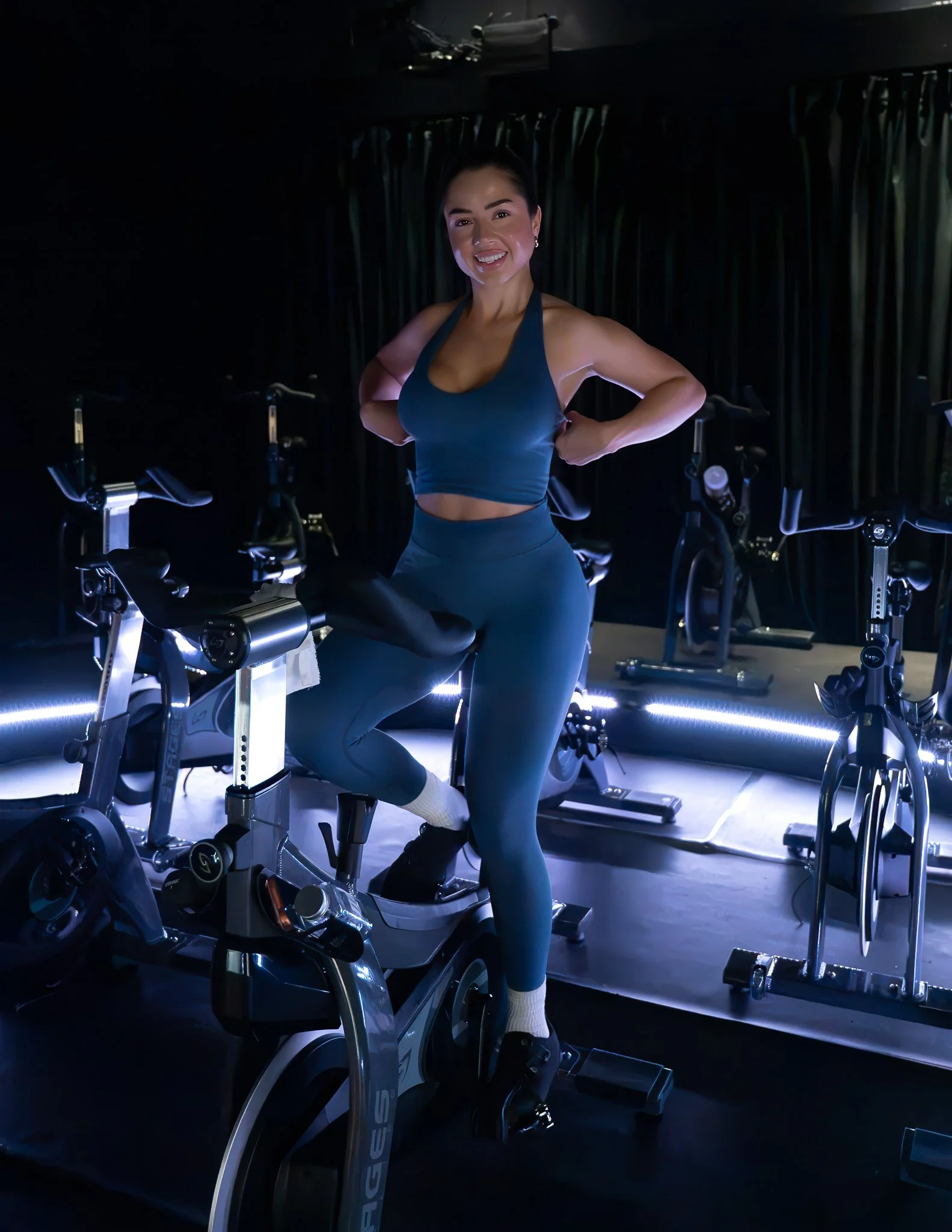 A woman smiling and standing on a stationary exercise bike in a dark fitness studio with other bikes and black curtains in the background.
