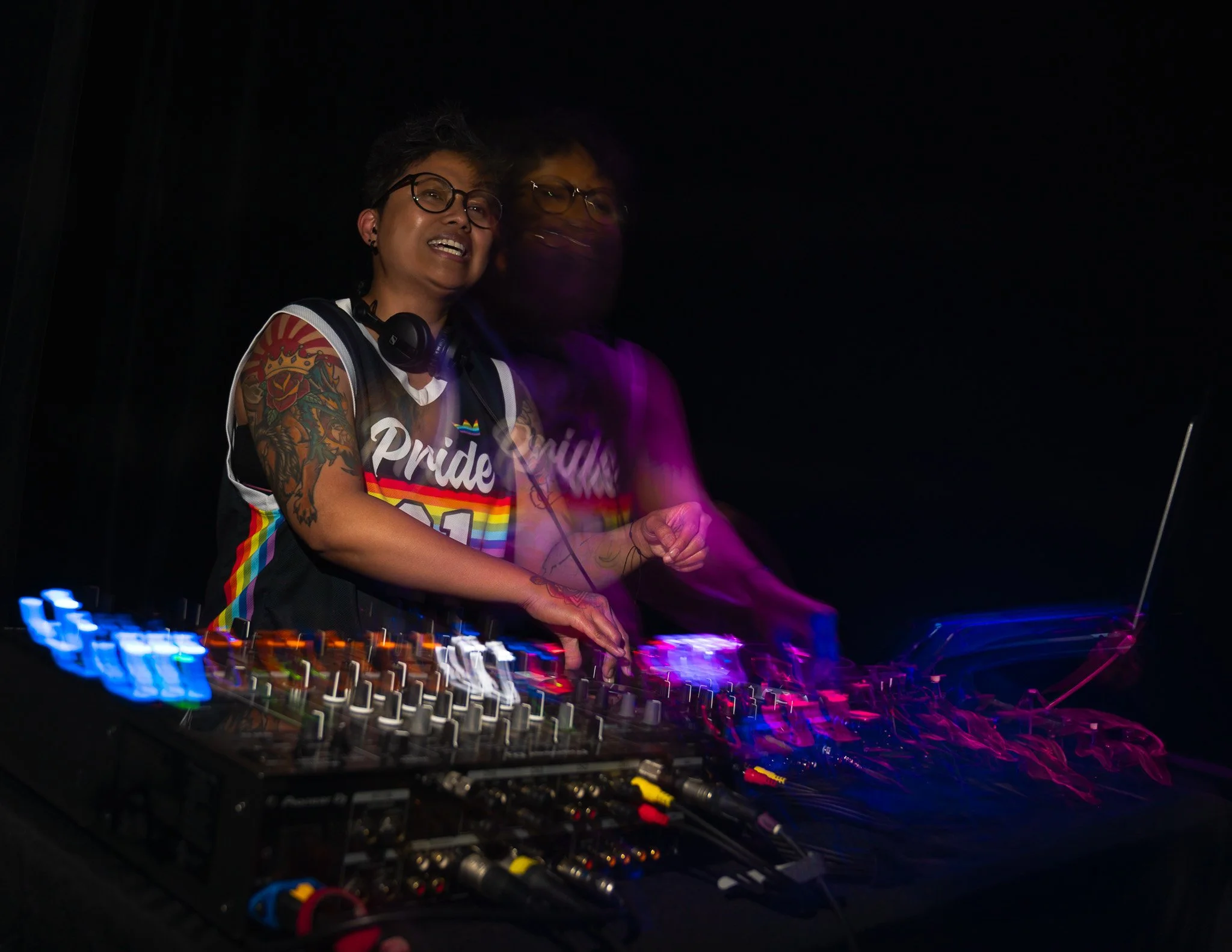 A DJ wearing glasses and a colorful Pride shirt is performing at a dark event, with equipment and colorful lights around.