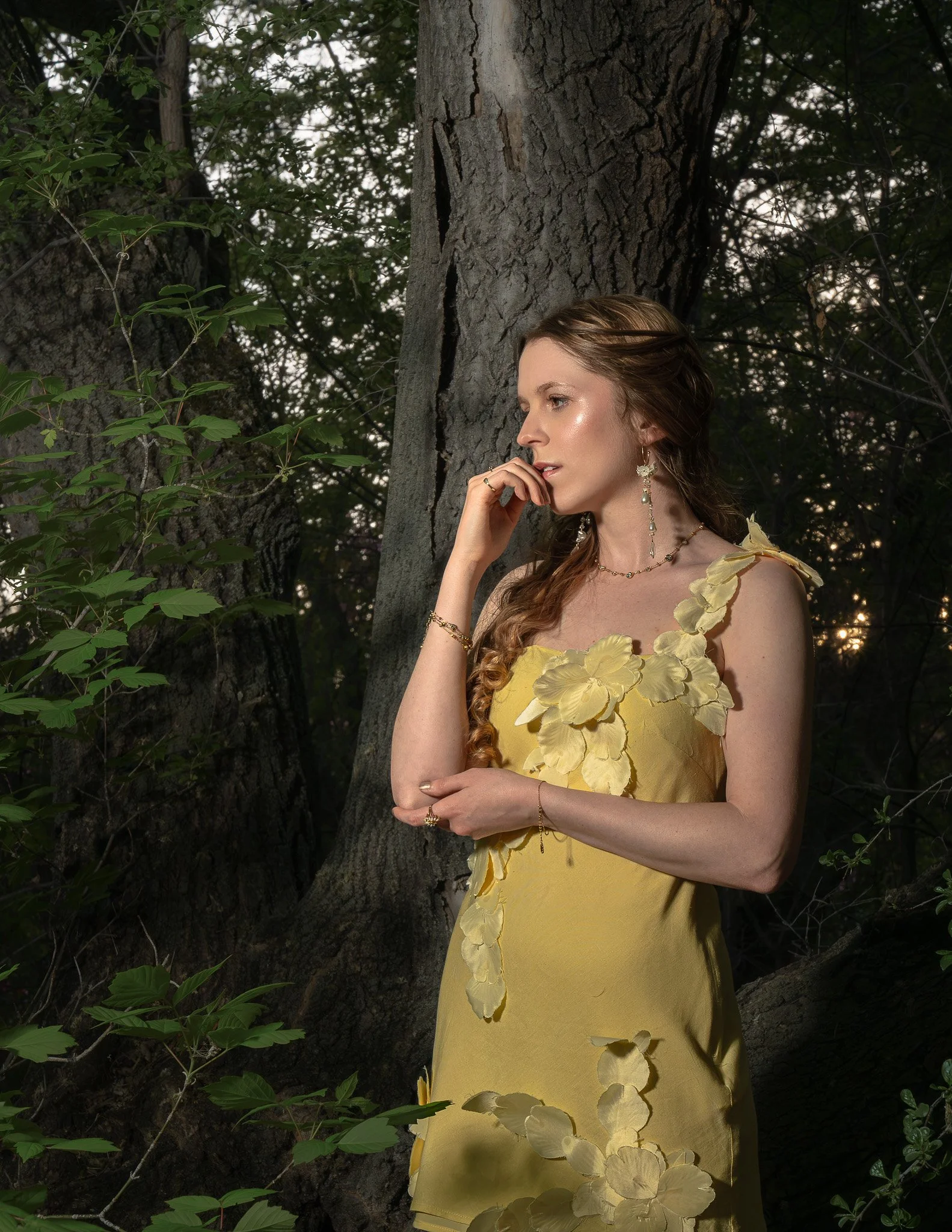 A woman wearing a yellow dress with leaf embellishments stands against a tree in a forest, with her hand near her face, surrounded by greenery and dappled sunlight.