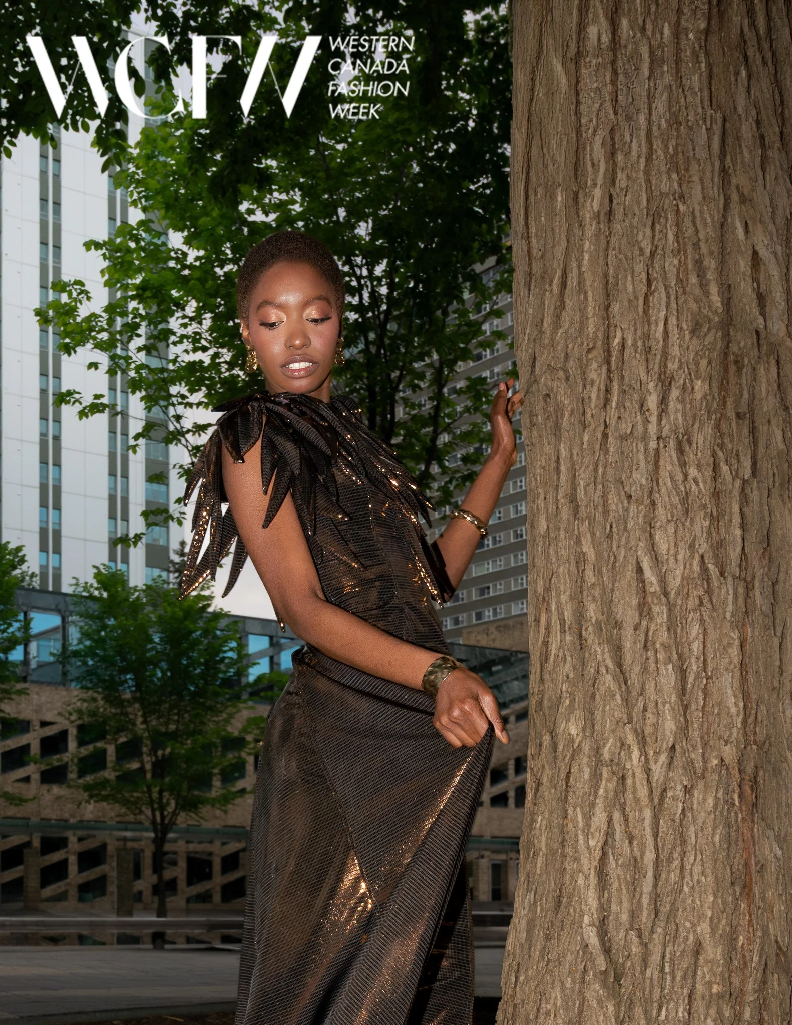 A woman dressed in a shiny black dress with shoulder bows is standing outdoors near a large tree, with modern buildings in the background, during the Western Canada Fashion Week.