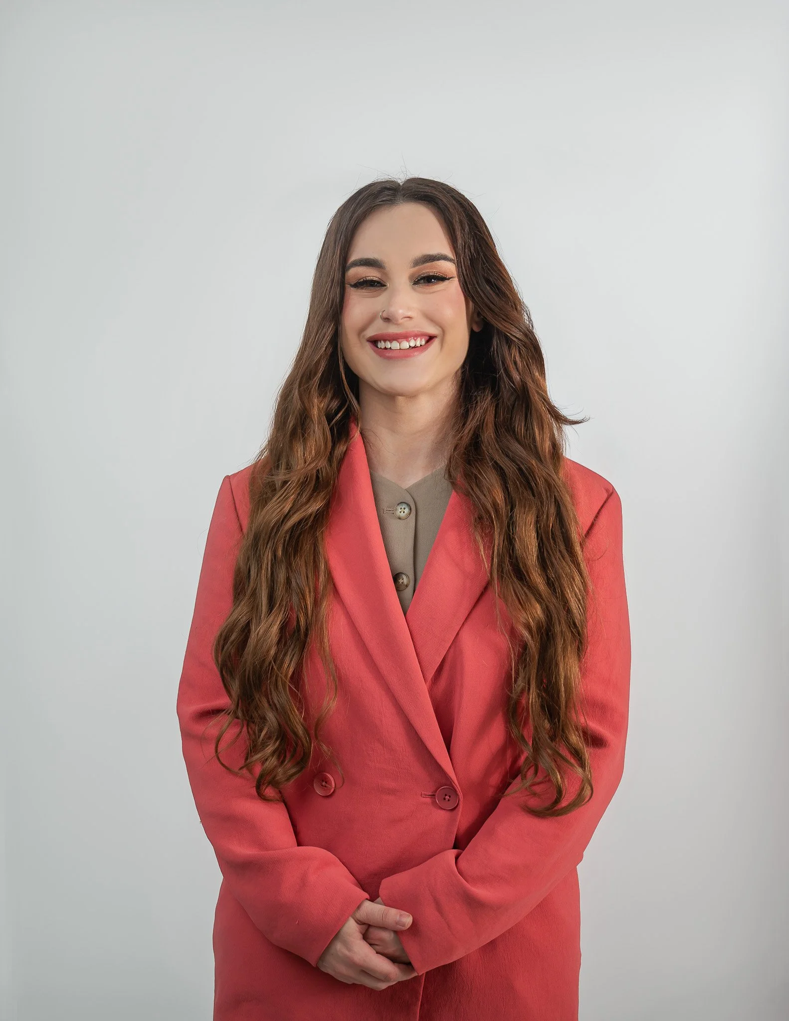 A young woman with long wavy hair and light skin, wearing a coral blazer over a beige top, smiling and standing against a plain light gray background.
