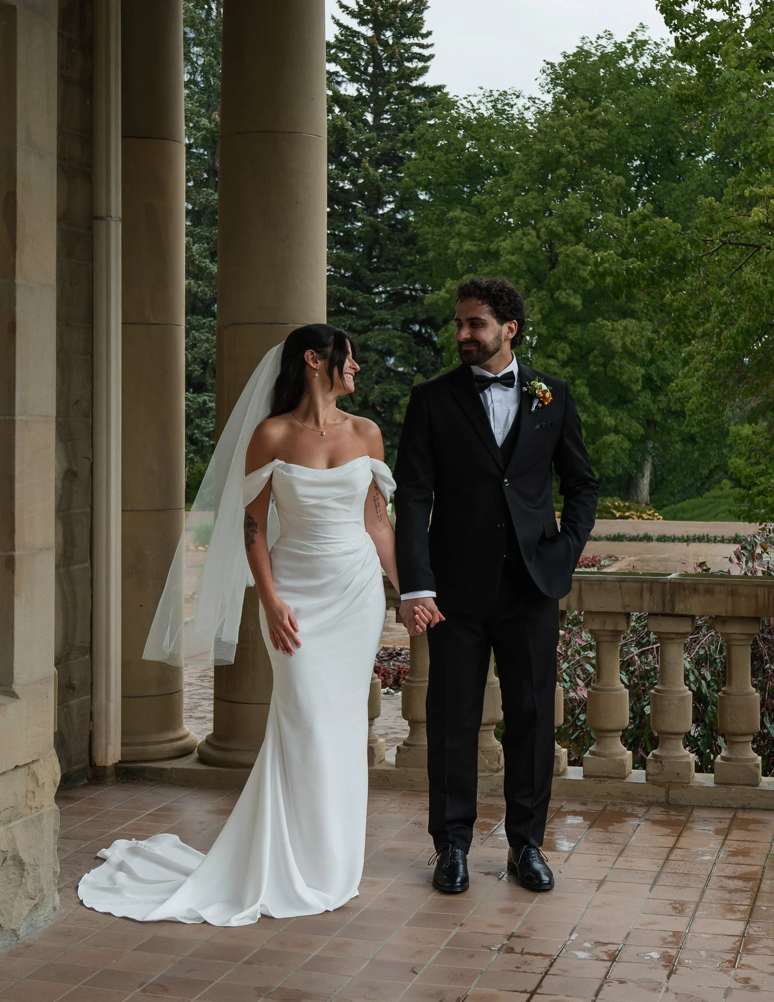 A bride and groom walking hand in hand outdoors on a wet tiled porch with large columns and green trees in the background. The bride is wearing a strapless white wedding gown and veil, and the groom is dressed in a black suit with a bow tie and bouto
