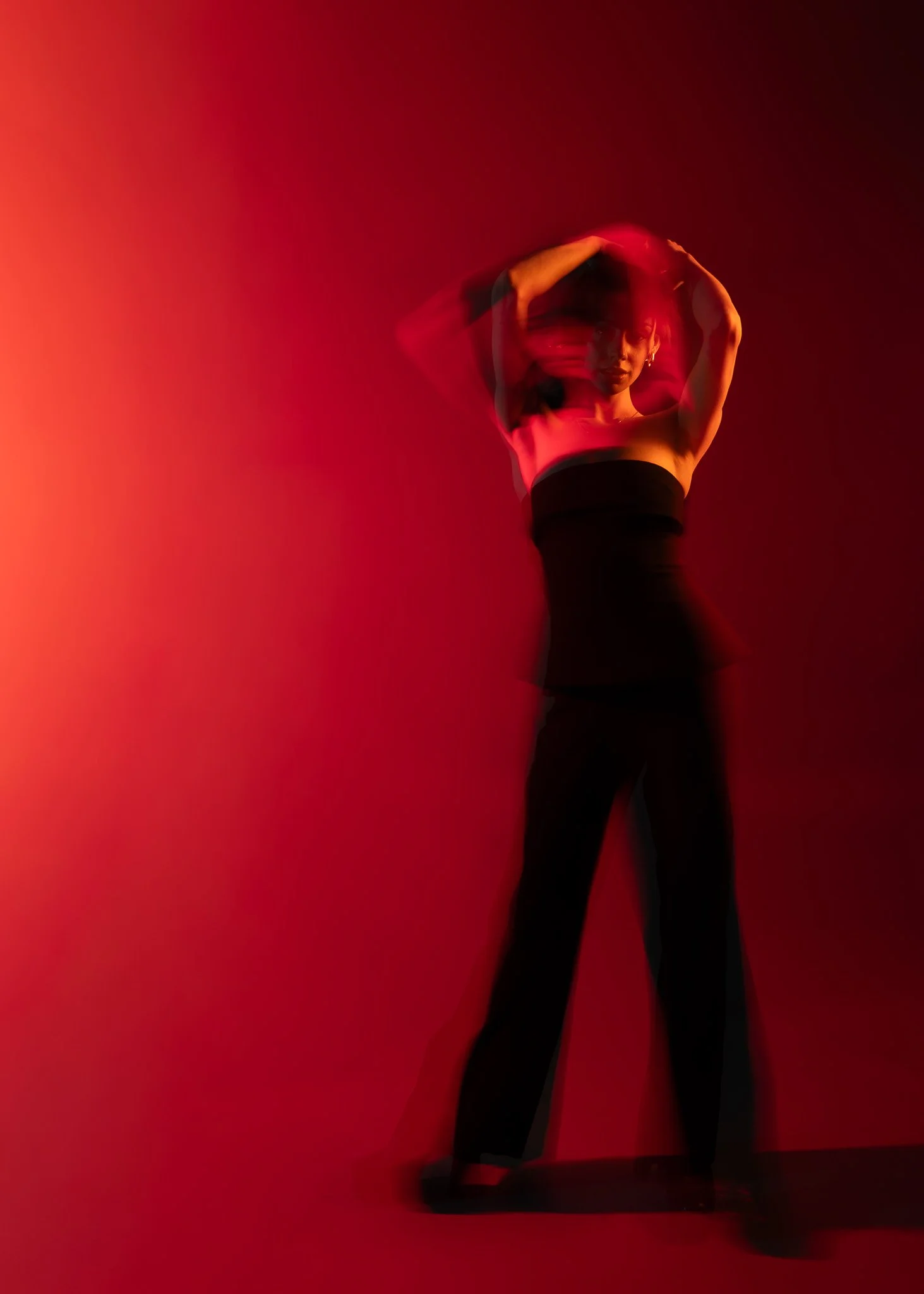 A woman standing against a red background, with her hands in her hair, wearing a black strapless top and black pants.