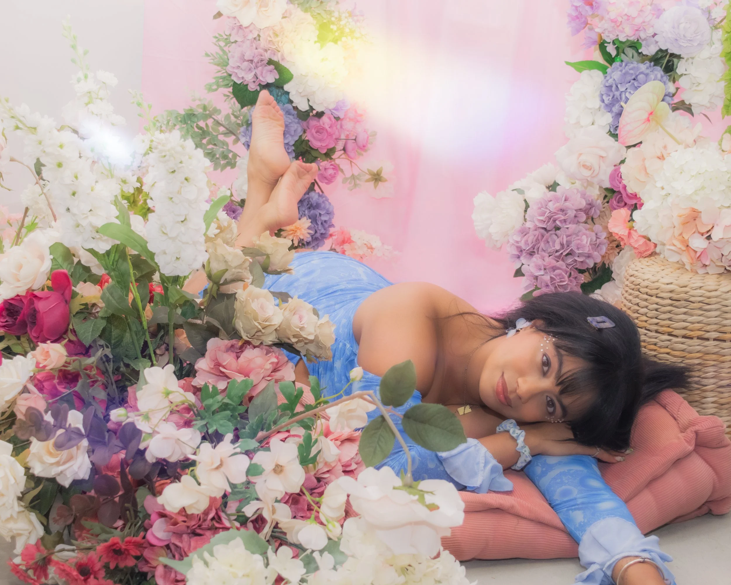 A young woman lying on the floor among colorful artificial flowers, surrounded by pink, purple, white, and peach blossoms, with a pink background.