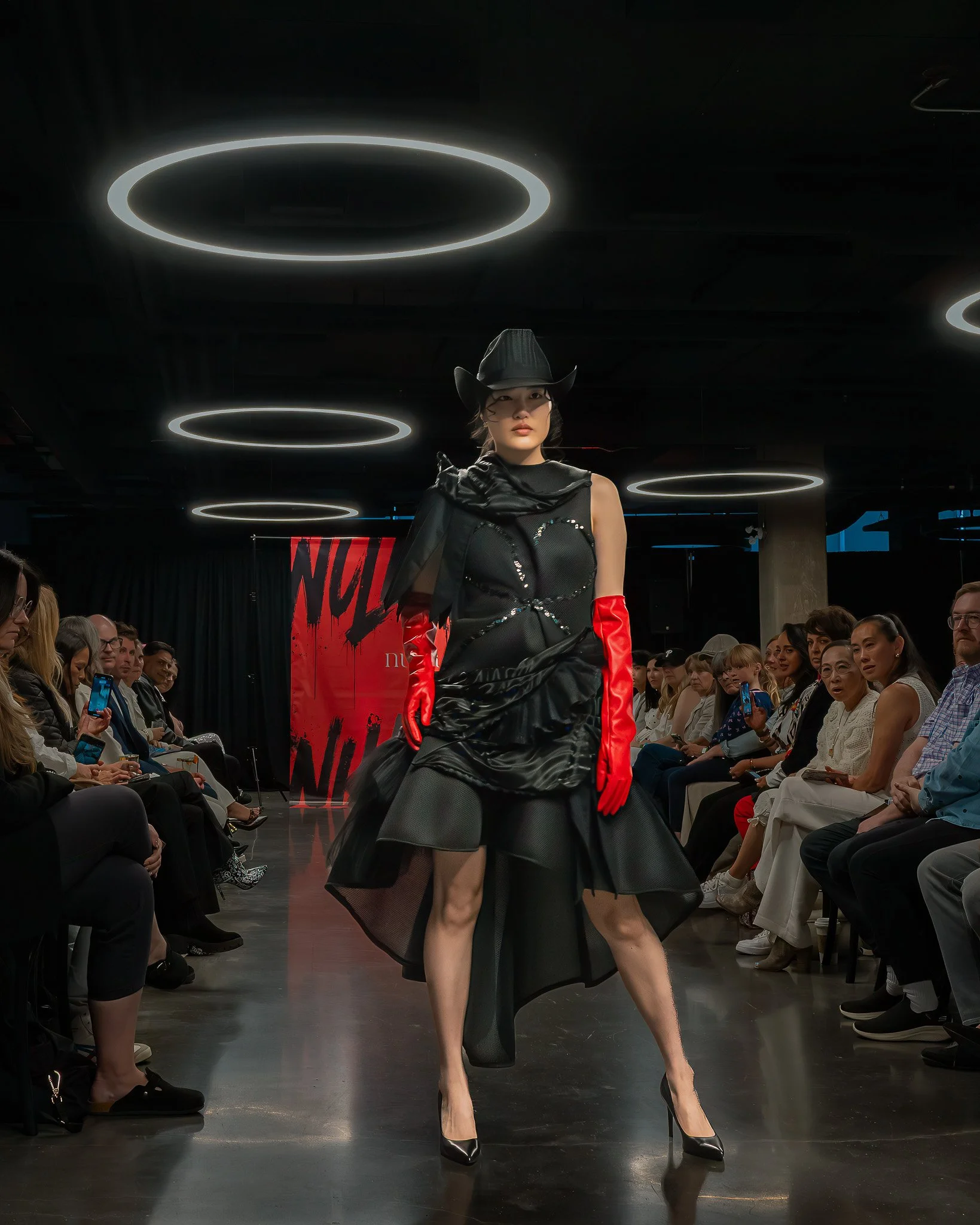 A model walking on a runway during a fashion show, wearing a black dress with a matching wide-brimmed hat, and red gloves, with an audience seated on either side watching the show.