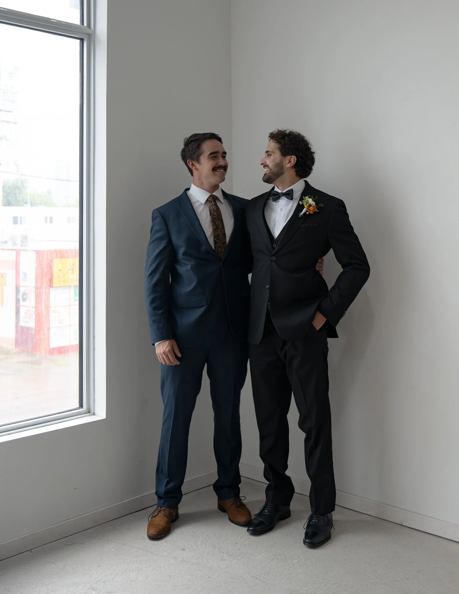 Two men in suits stand close together, smiling and looking at each other, next to a large window in a bright room.