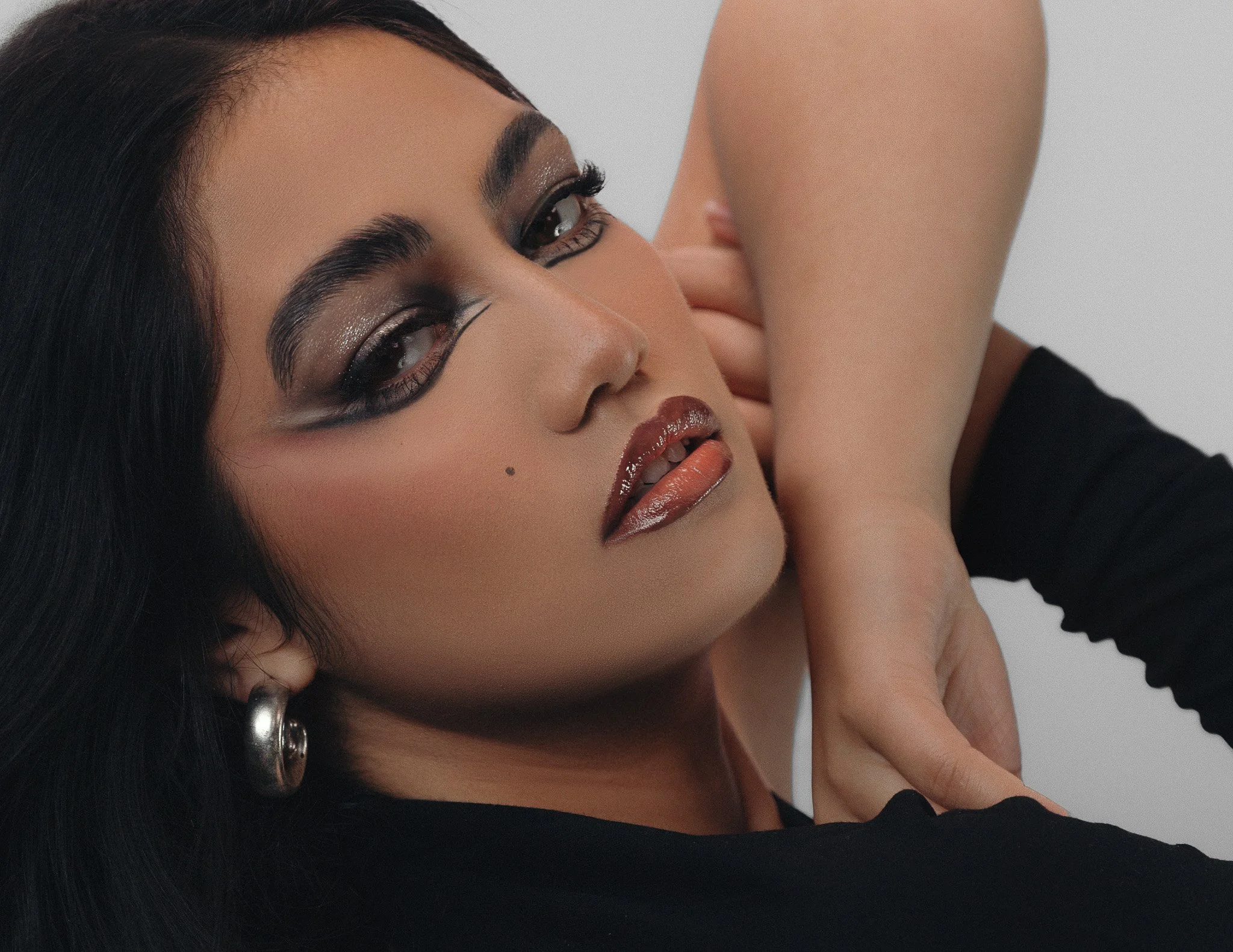 Close-up of a woman with dark hair and makeup, wearing hoop earrings and a black top, with her arm raised.