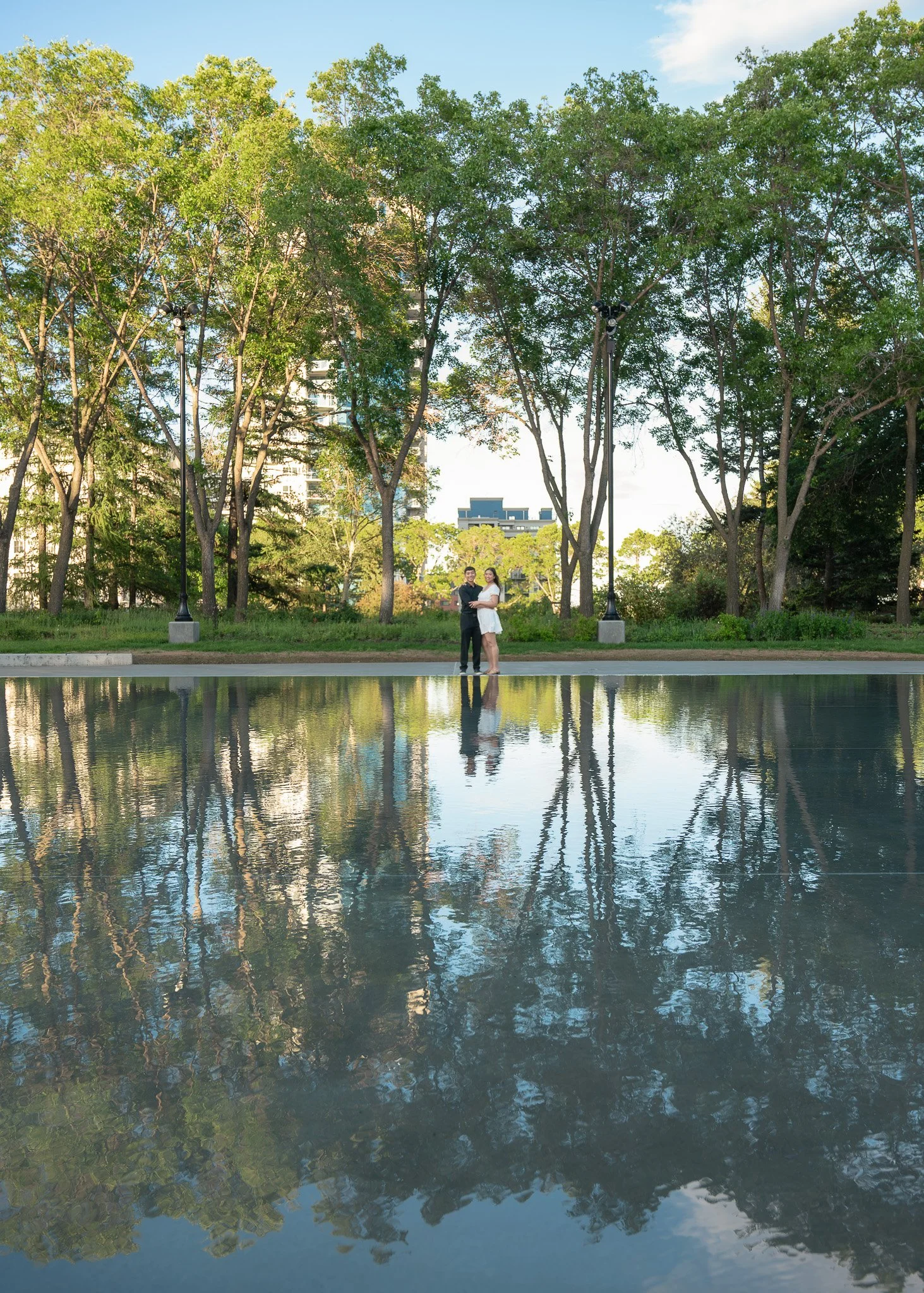 A couple standing in a park by a pond with green trees and modern buildings in the background, reflecting in the water.