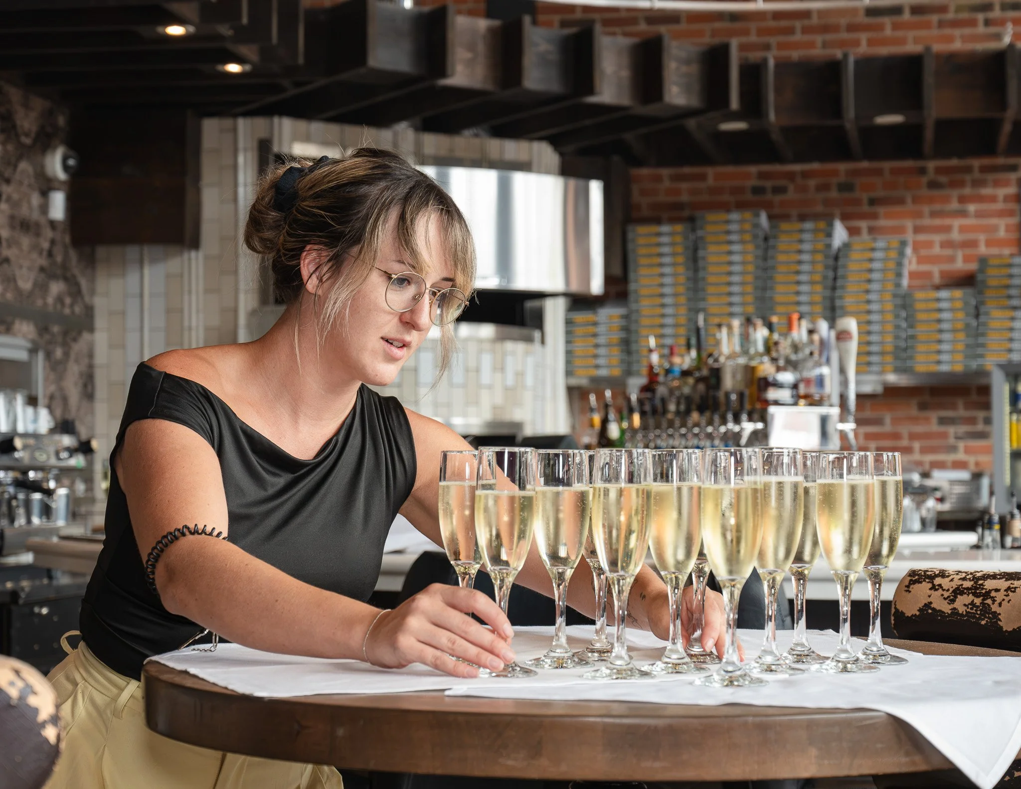 A woman with glasses arranging champagne glasses on a table in a restaurant or bar setting.