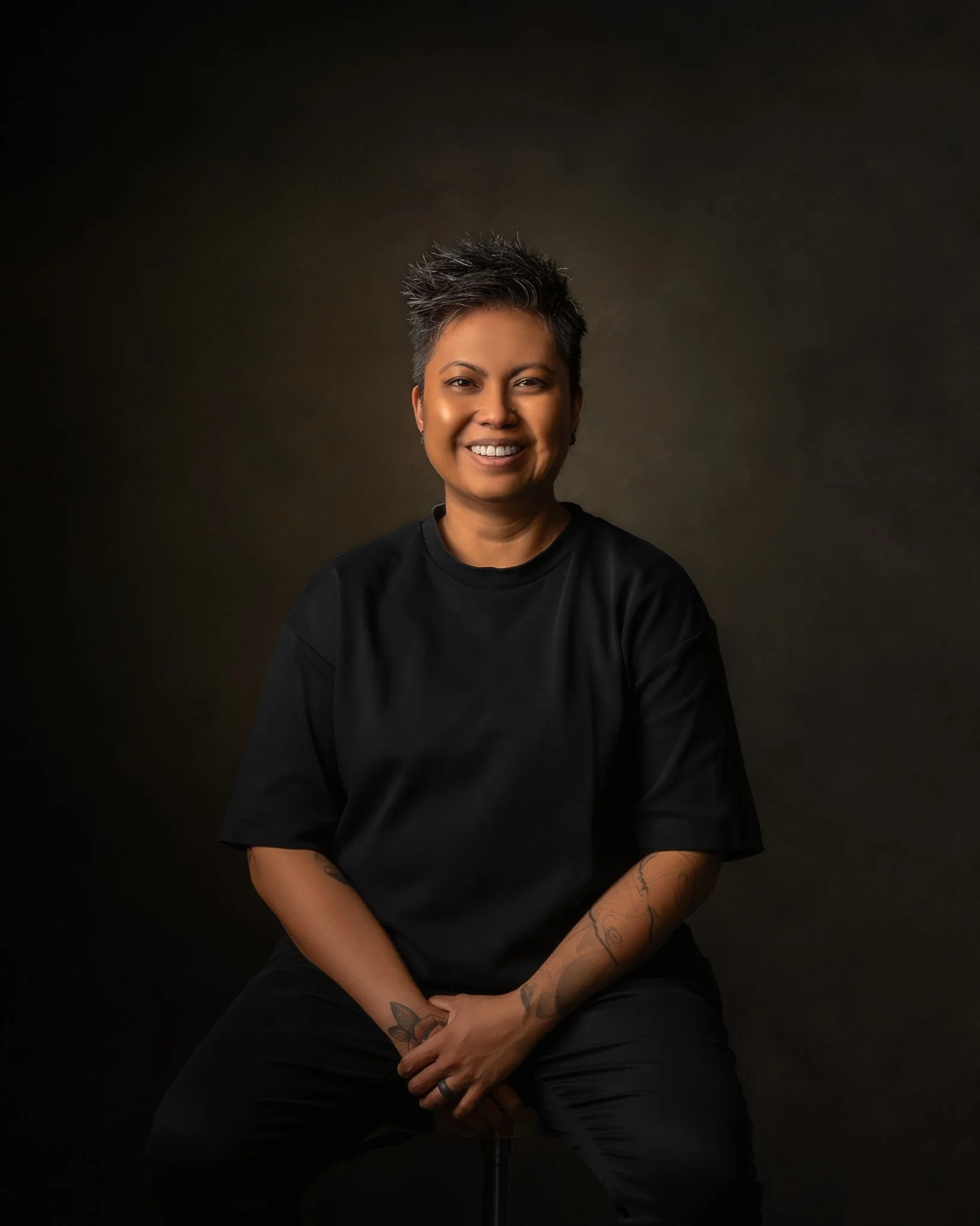 Portrait of a woman with short, spiked gray hair, smiling, sitting against a dark background, wearing a black shirt, with visible tattoos on her arms.