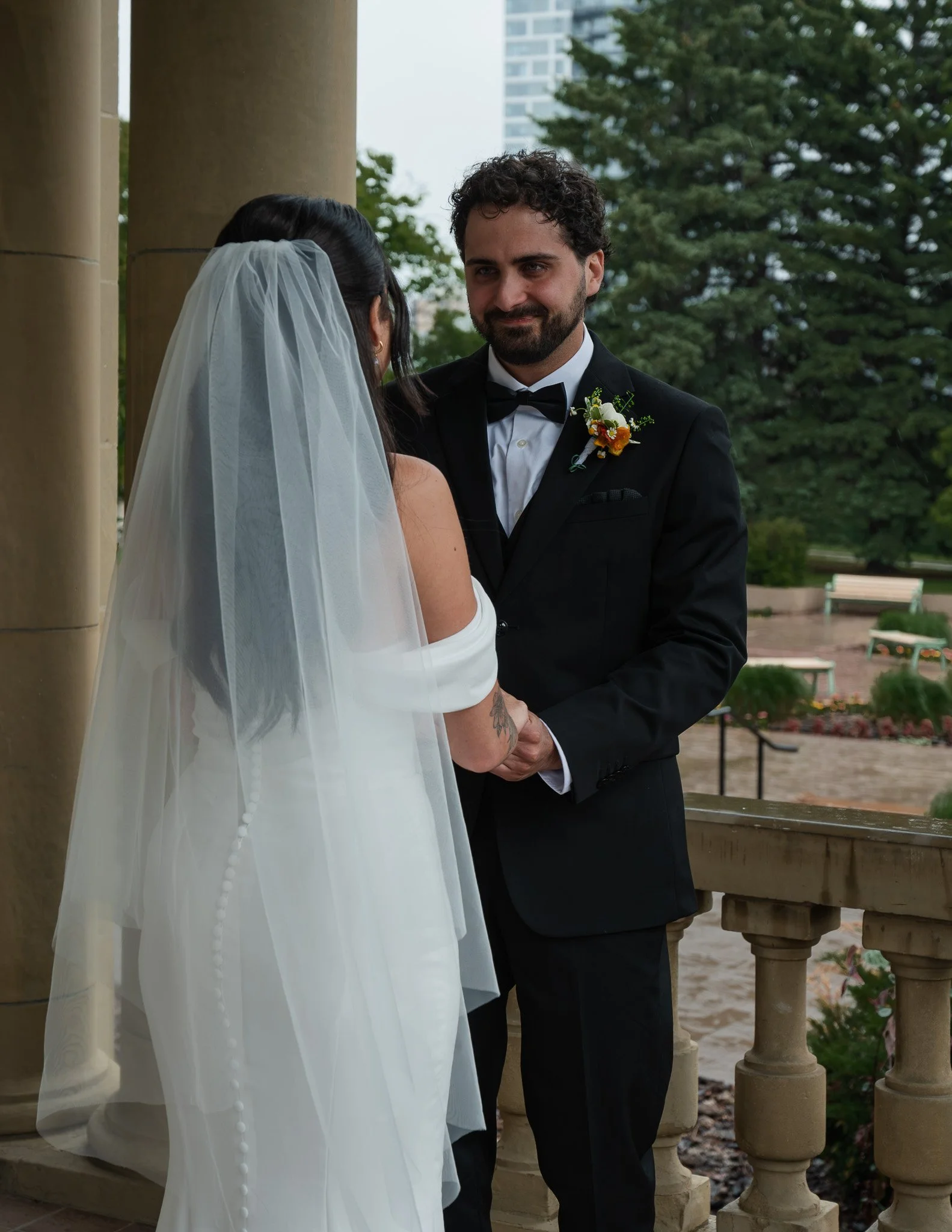 A groom in a black tuxedo with a boutonniere, looking at a bride with a veil, during a wedding ceremony outdoors in front of green trees and a stone railing.