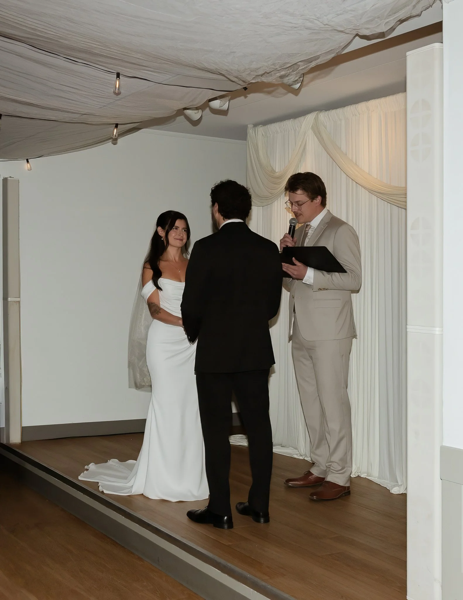A wedding ceremony with three people standing on a wooden stage with a white curtain backdrop, the bride in a white gown, the officiant in a beige suit, and the groom in a black suit, during the daytime, with decorative drapes and string lights overh