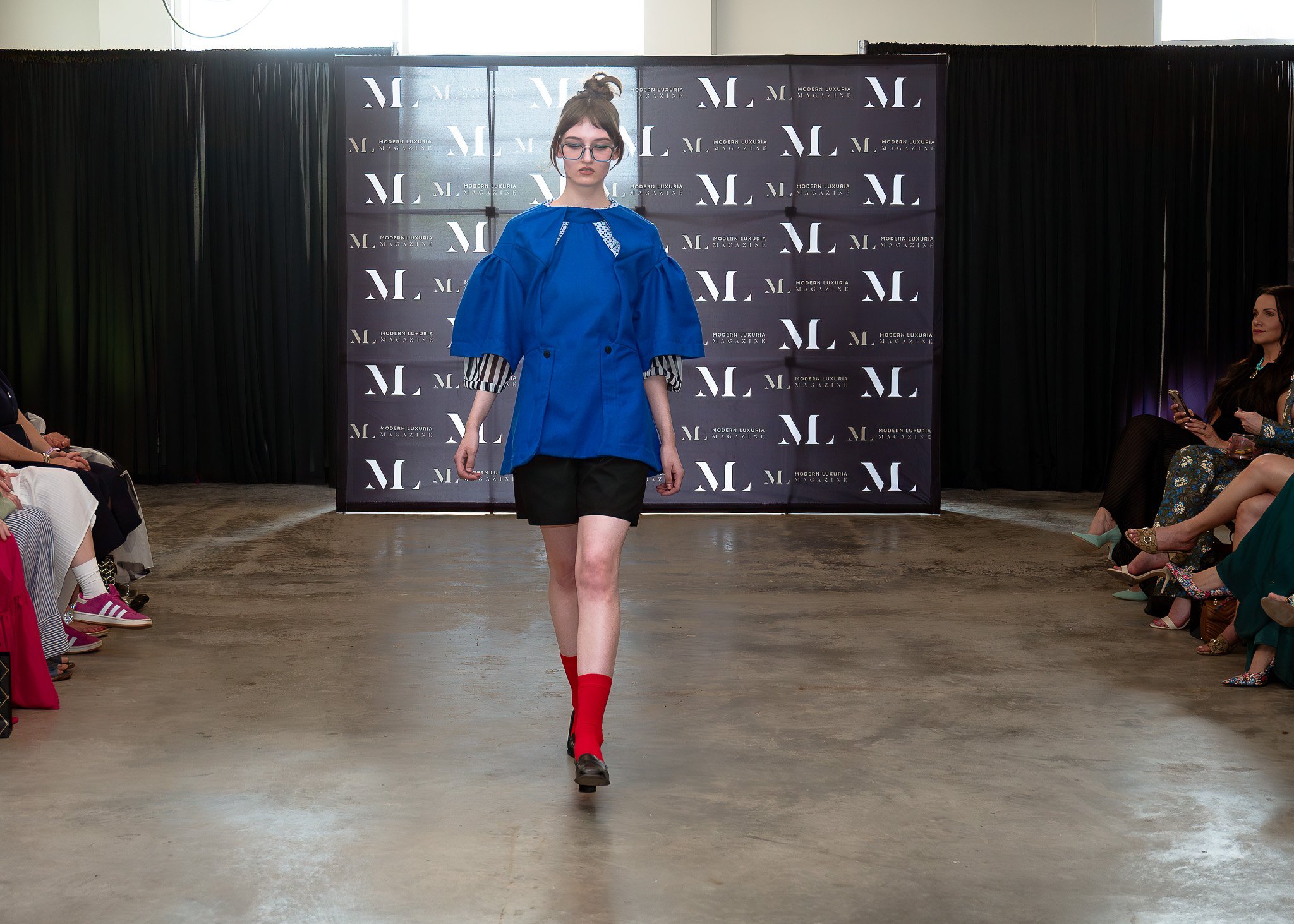 A female model walks down the runway during a fashion show. She is wearing a blue jacket with striped sleeves, black shorts, and bright red socks with black shoes. The background features a black curtain and a banner with the logo 'MODERN LUXURIA MAG