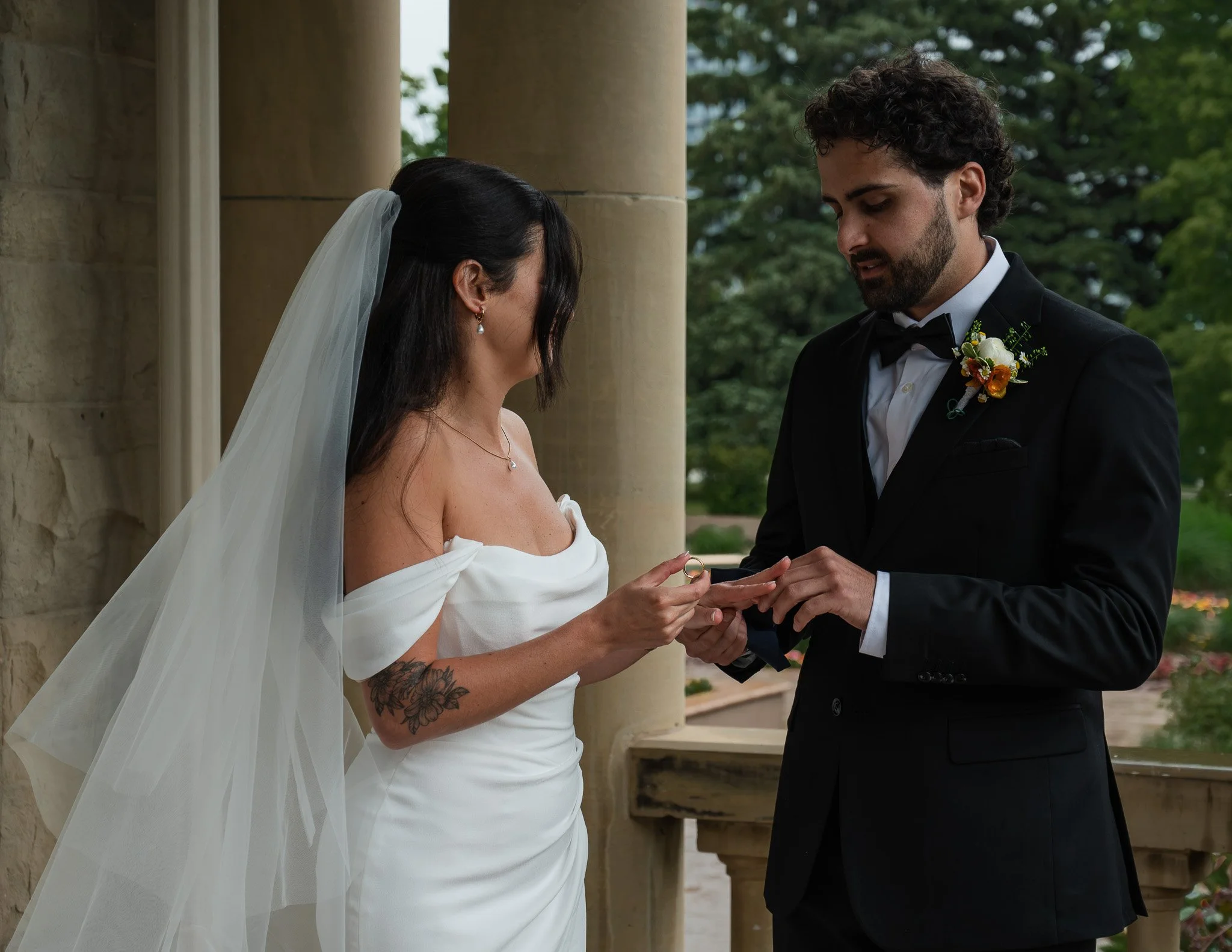 A bride and groom exchanging rings outside during a wedding ceremony, with the bride wearing a white dress and the groom in a black tuxedo, standing under a stone column with greenery in the background.