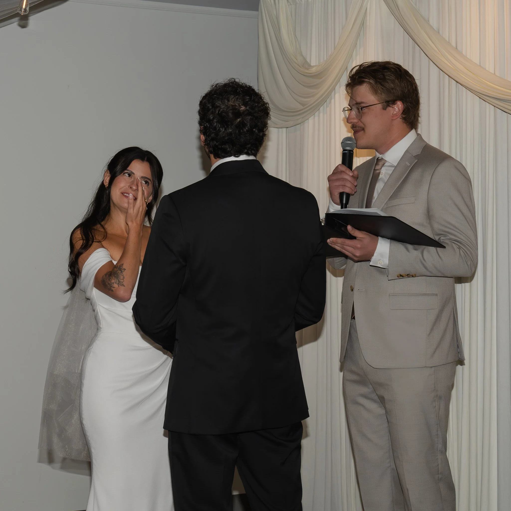 A wedding ceremony with a woman in a white dress and a man in a light gray suit standing in front of a curtain, with another person in black between them.