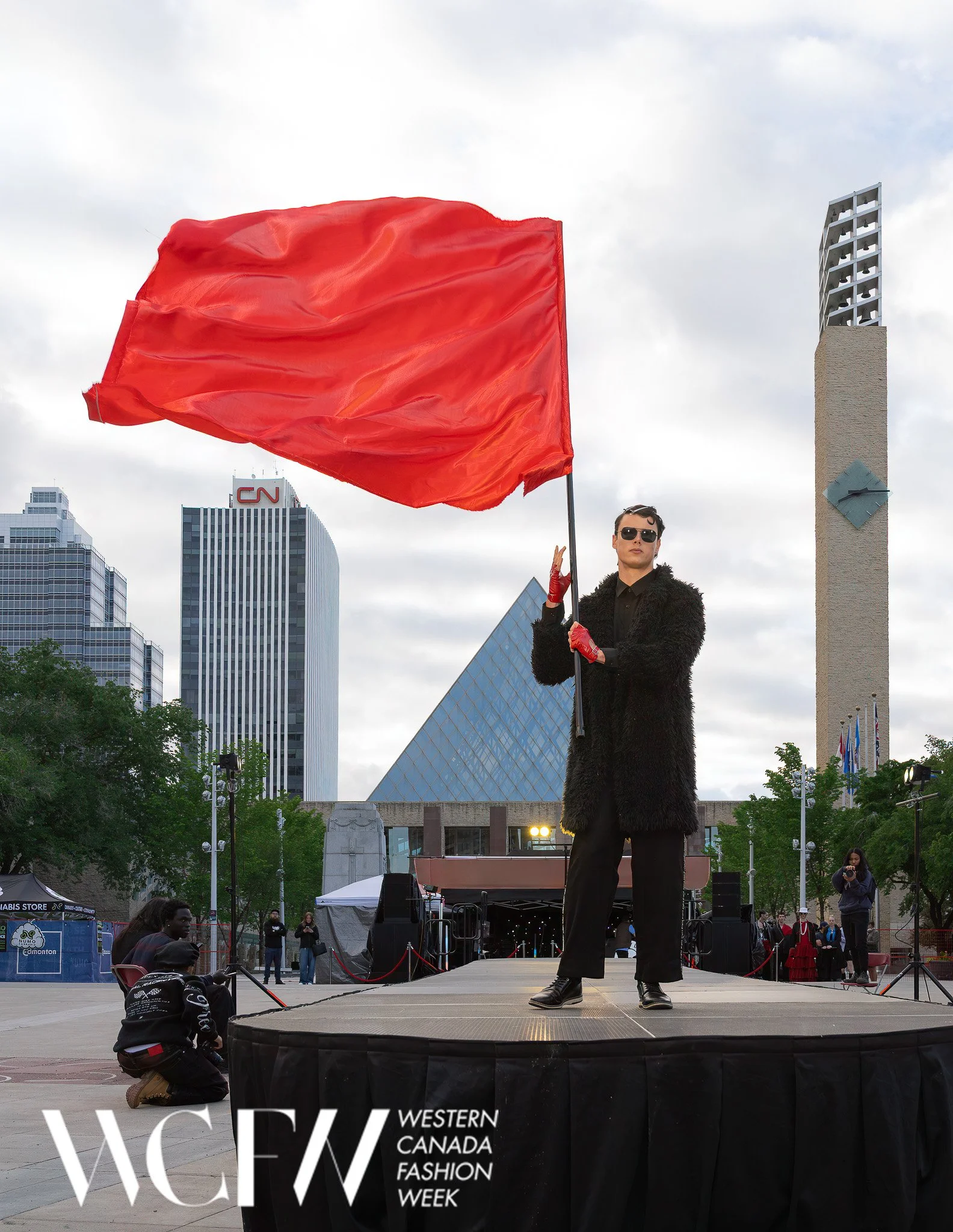 A person dressed in black with sunglasses is standing on a stage holding a red flag. The background shows a cityscape with tall buildings, some with logos, and a cloudy sky. There are several people and photographers in front of the stage, and a bann