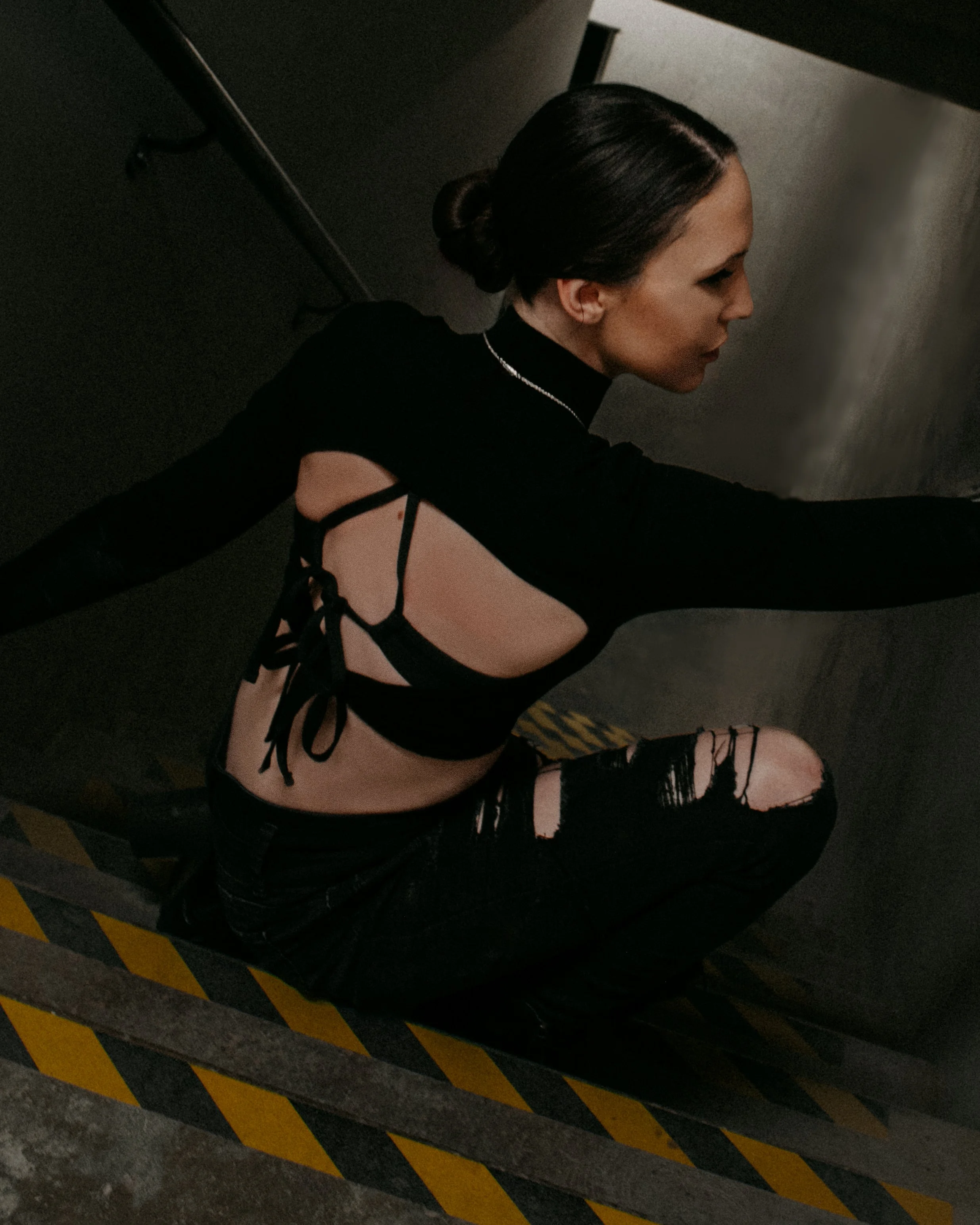 A woman crouching against a wall in a dark setting, wearing a black top with an open back and ripped black jeans.