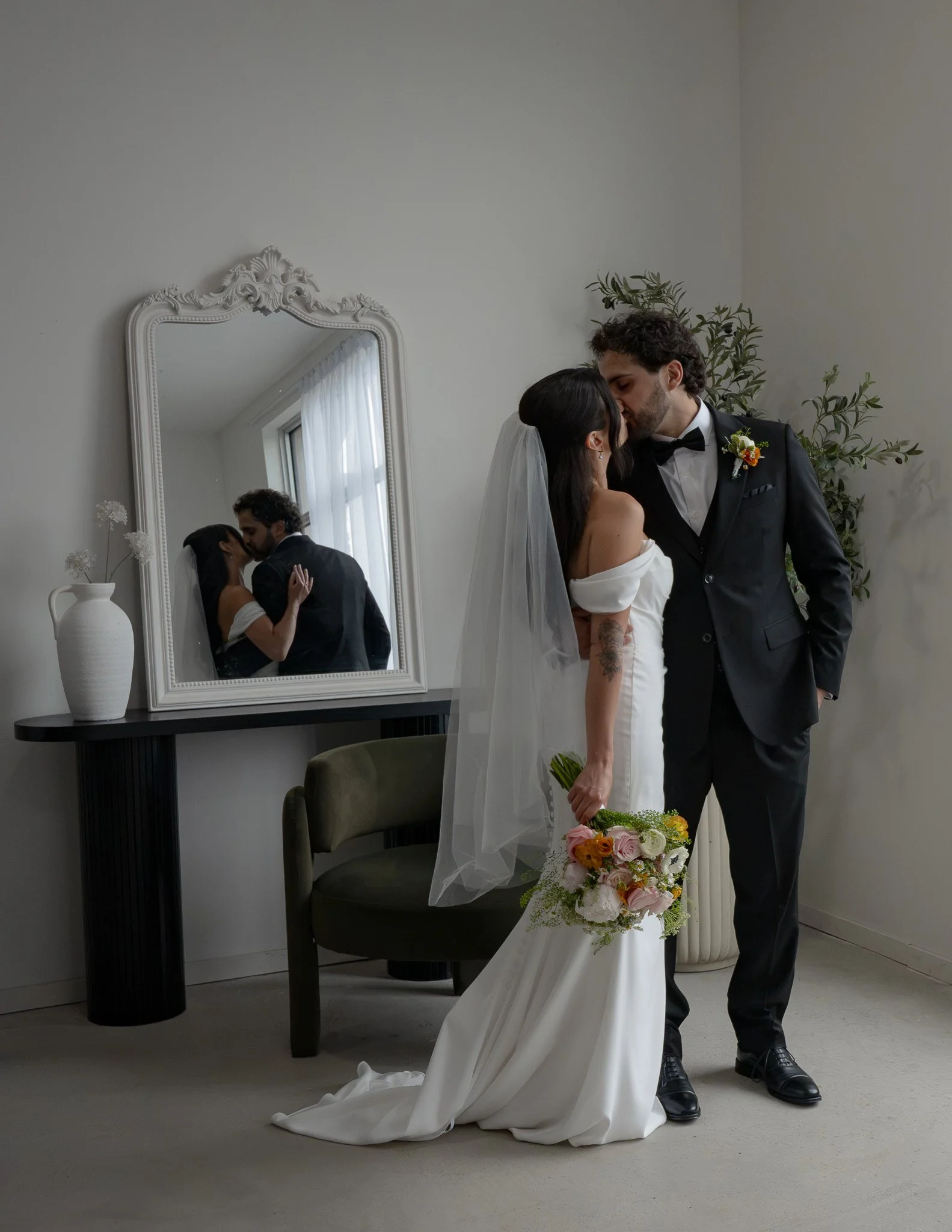 A bride and groom share a kiss in front of a large mirror in a room with white walls and natural light. The bride is holding a bouquet of flowers, and the groom is dressed in a black tuxedo with a boutonniere.
