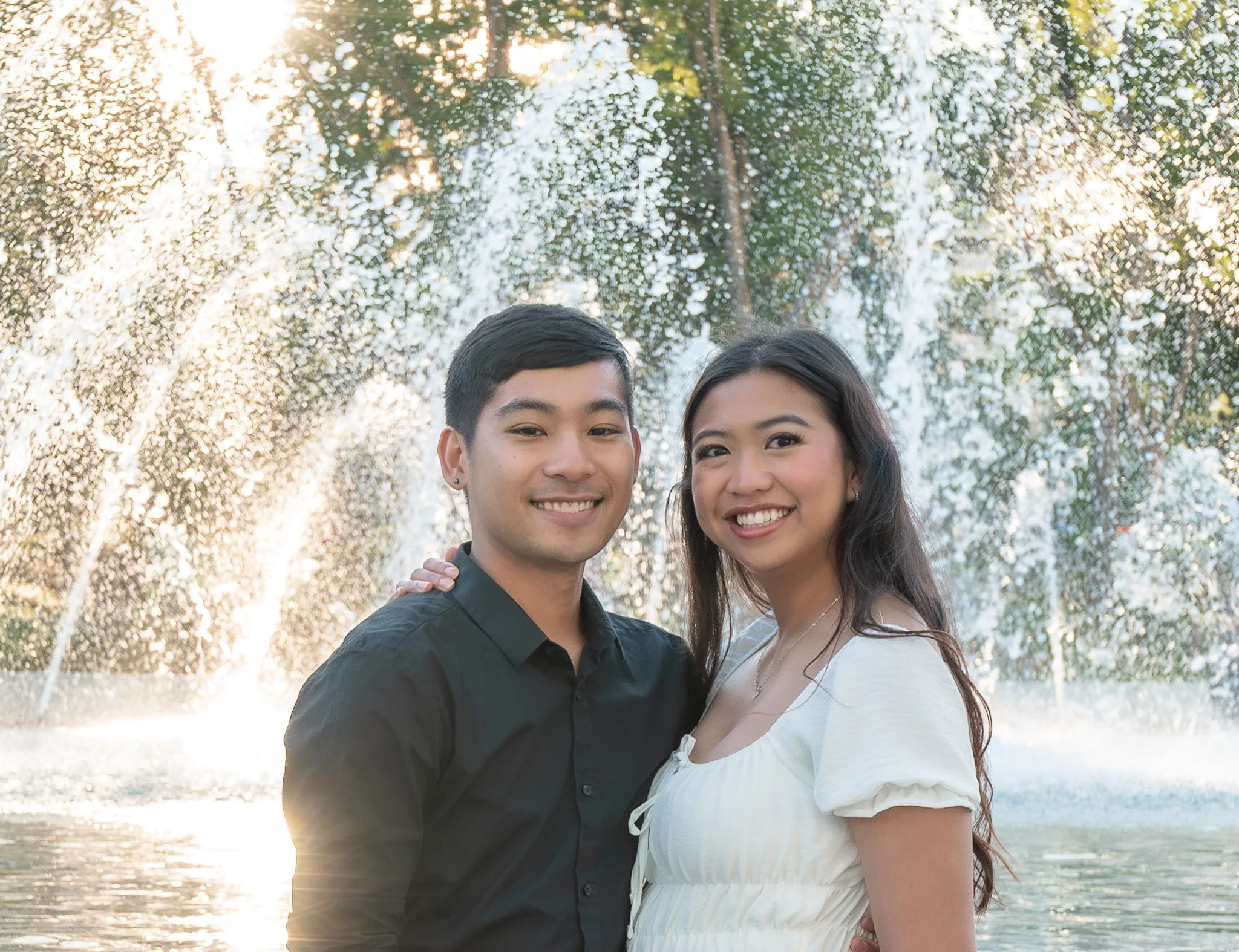 A happy young couple taking a selfie outdoors in a park during sunlight, with trees in the background.