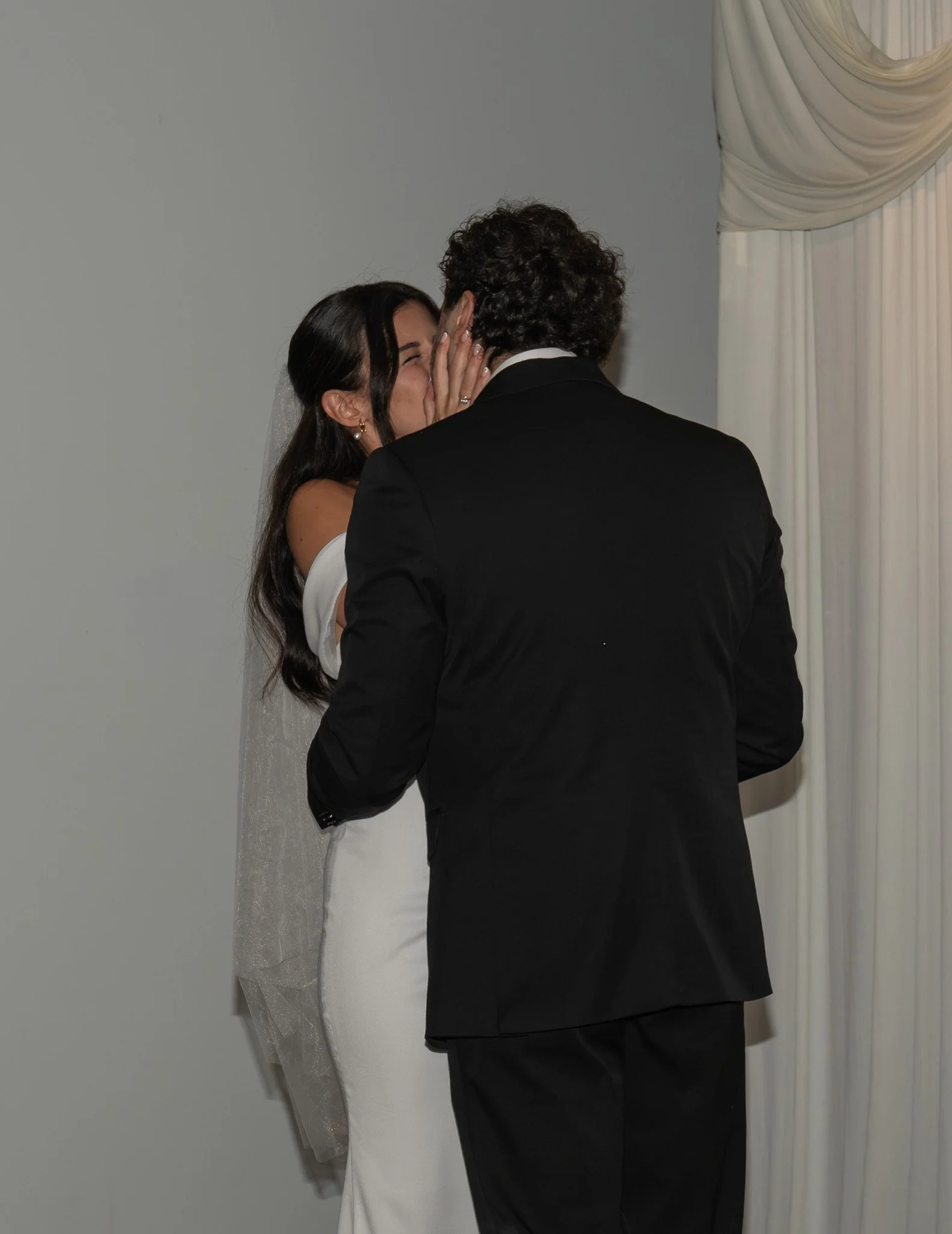 A bride and groom are sharing a kiss during their wedding ceremony in front of a white curtain.