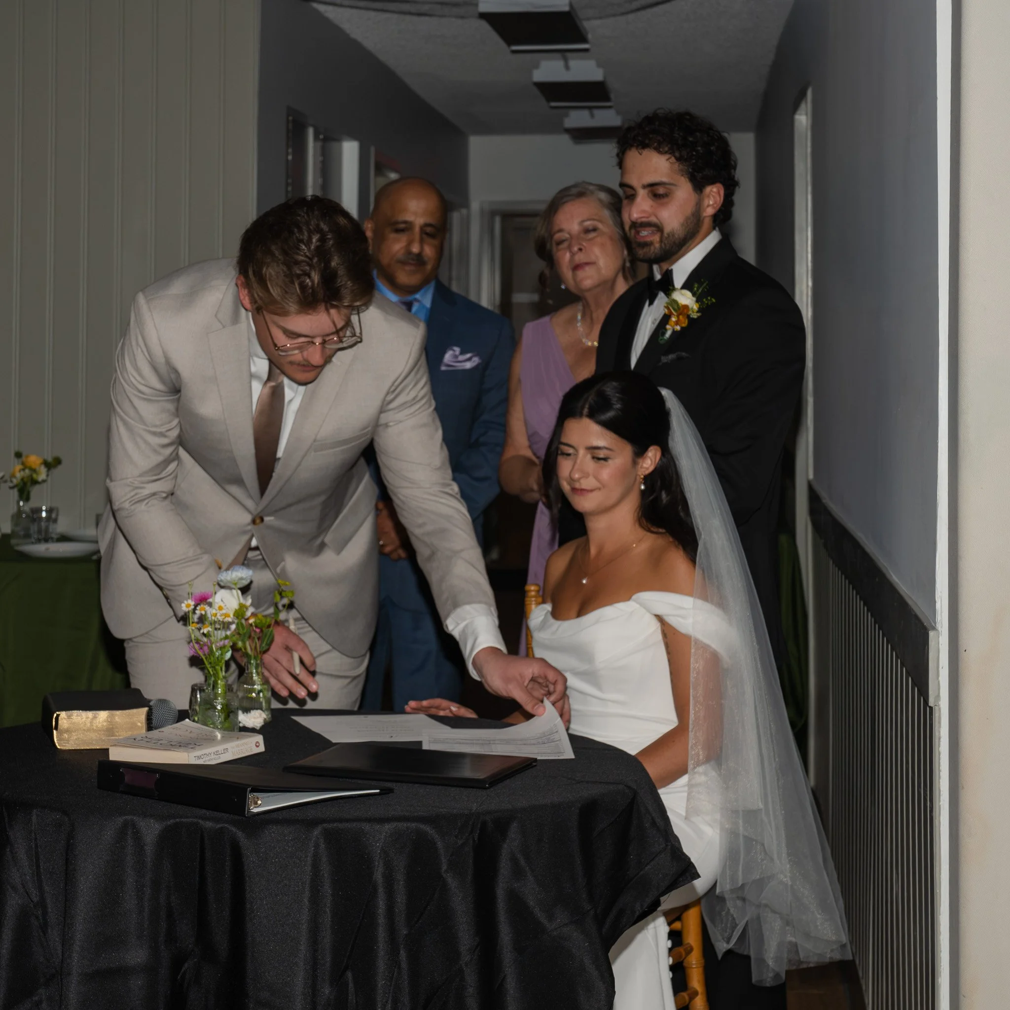 A group of people dressed formally standing in a dimly lit room, with some looking at a paper or document on a table covered with a black cloth, which also has a small glass and a book. The person in the cream-colored suit is signing or writing on th