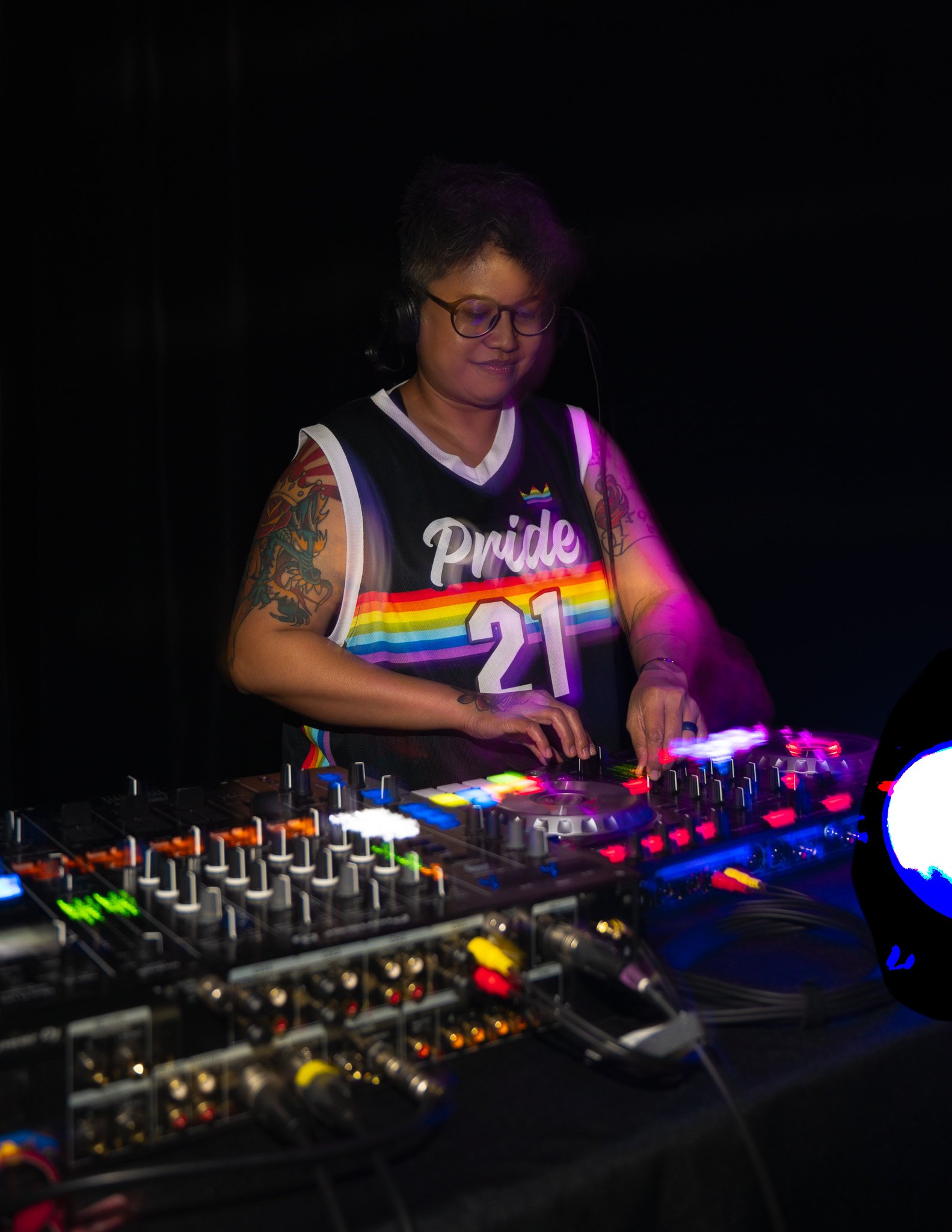 A DJ performing at a nightclub, wearing a Pride shirt with rainbow colors and the number 21, surrounded by DJ equipment with colorful lights.