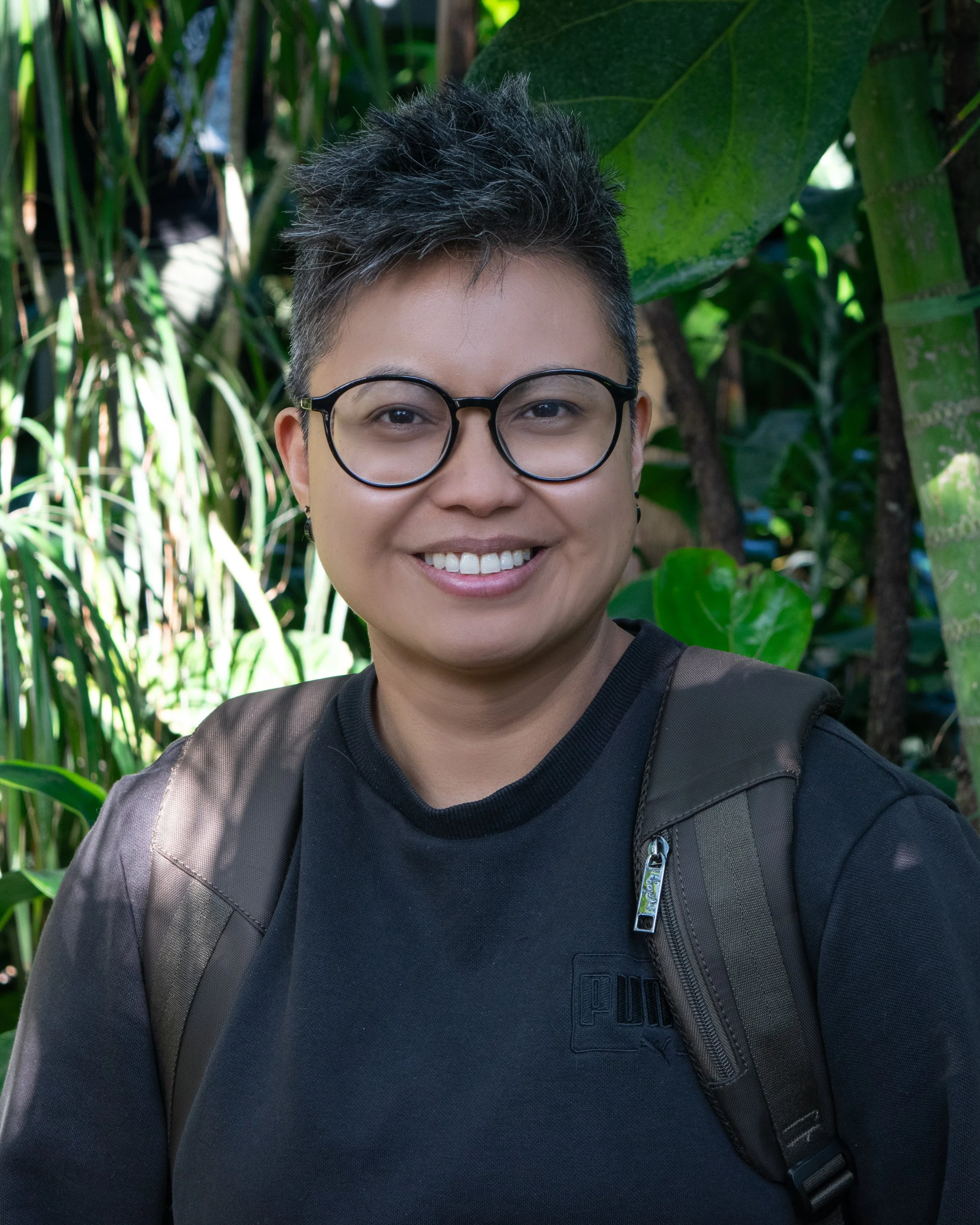 A smiling person with short dark hair, glasses, wearing a black Puma shirt, and carrying a backpack in a lush green jungle environment.