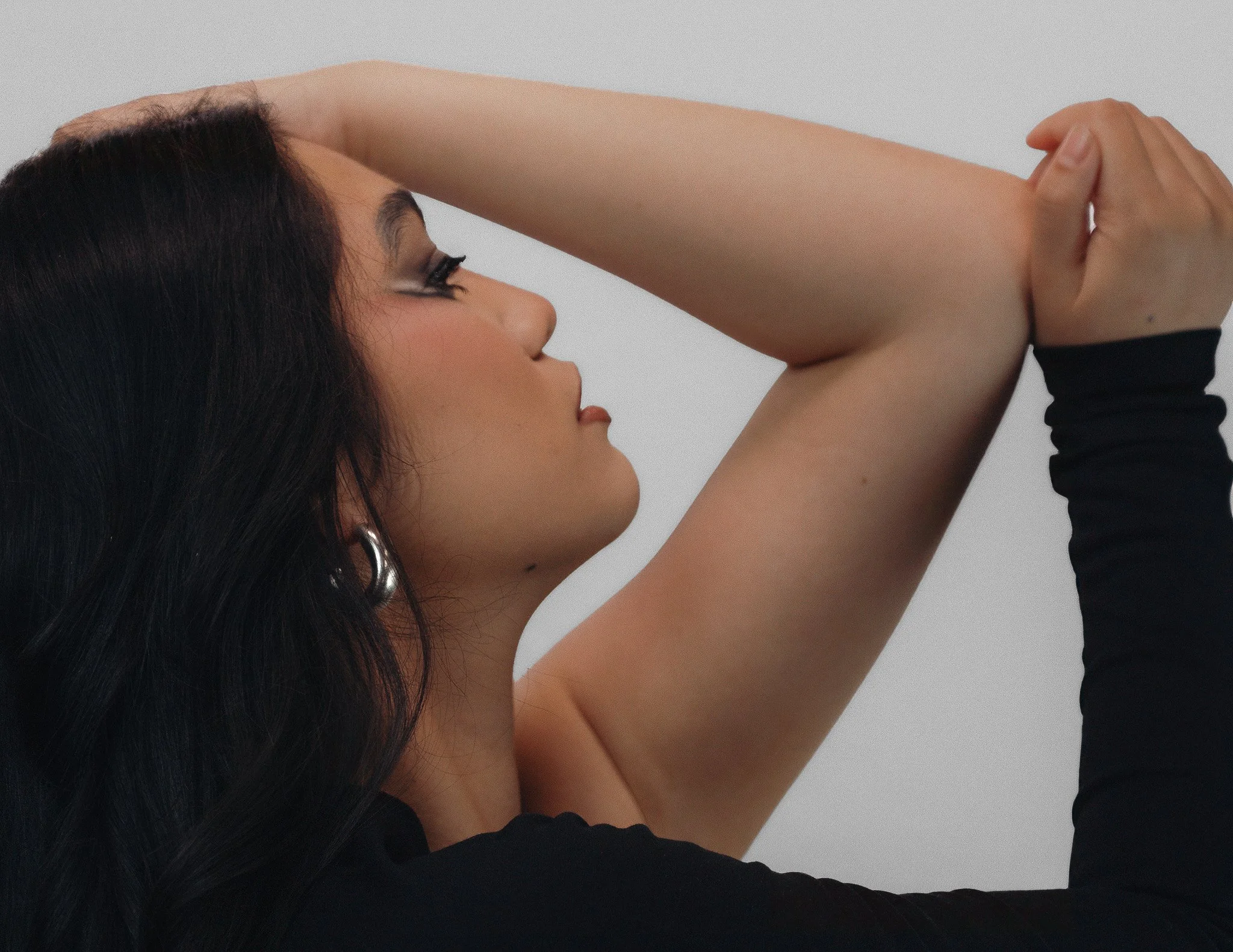 A woman with long dark hair and makeup, holding her arm above her head, wearing a black long sleeve top.