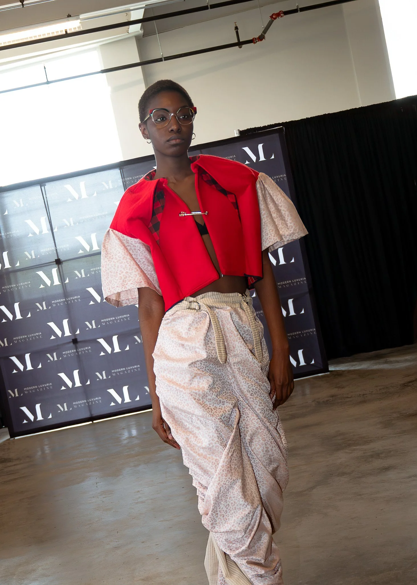 An individual with dark skin, short hair, glasses, and earrings, standing indoors in front of a backdrop with the words 'Modern Luxuria Magazine' repeated on it. They are wearing a layered outfit with a red top, patterned fabric, and beige patterned 