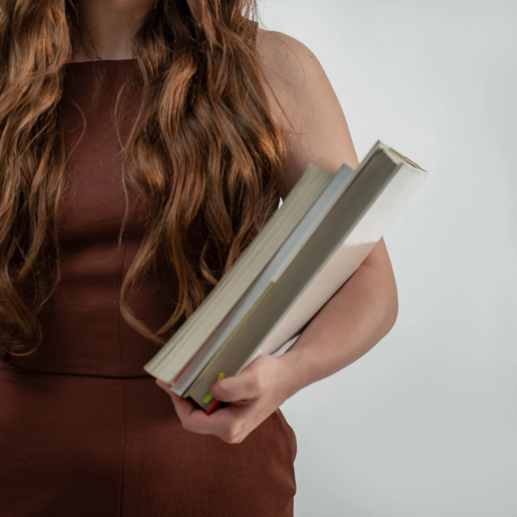 A woman with long, wavy red hair is holding a stack of books in her arms, wearing a sleeveless top, standing against a plain white wall.
