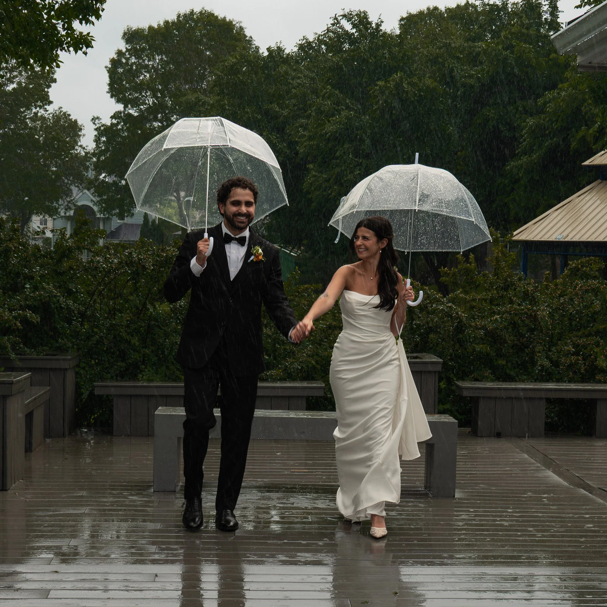 A man in a tuxedo and a woman in a wedding dress holding hands and walking outdoors in the rain, each holding a clear umbrella, with trees and houses visible in the background.