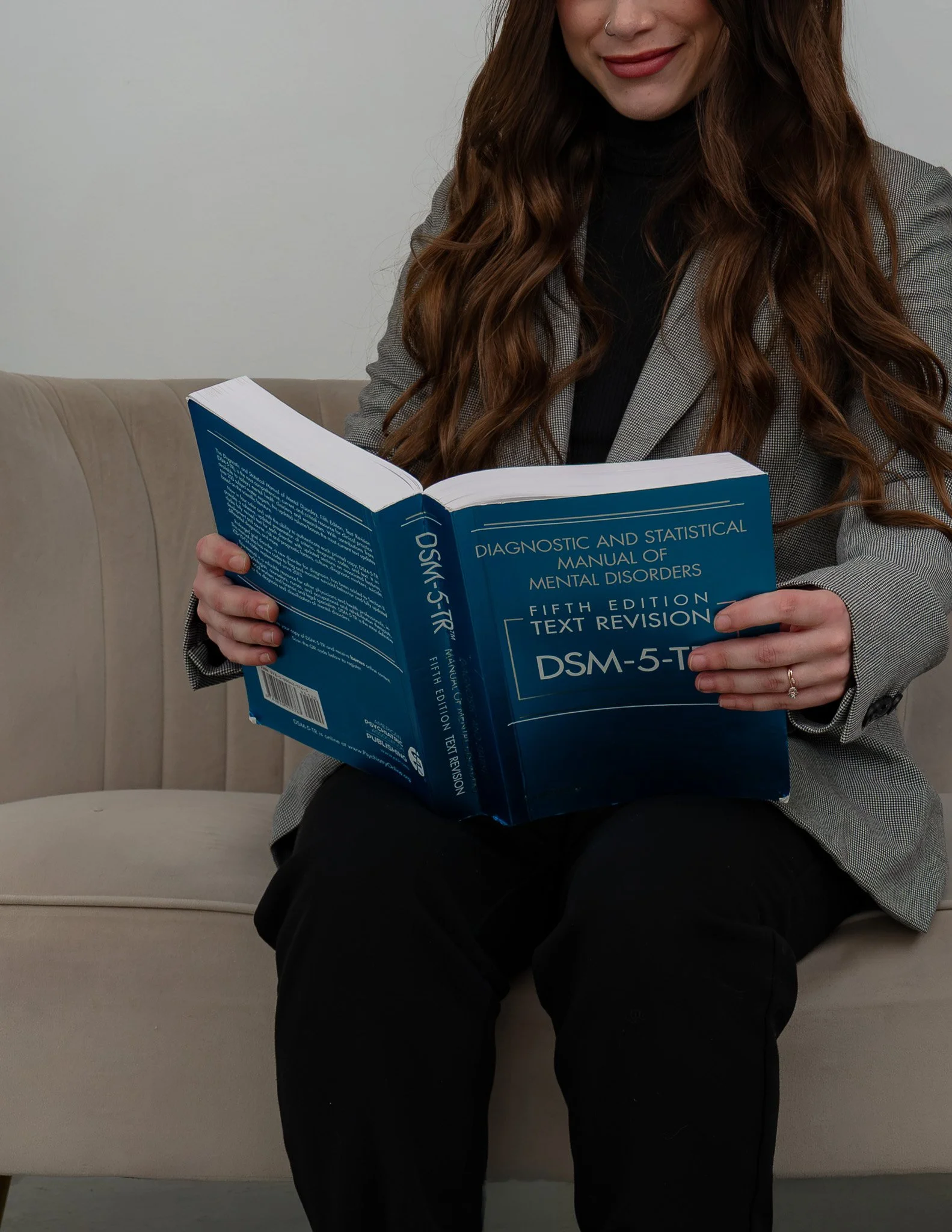 A woman with long, wavy brown hair, smiling slightly, sitting on a beige couch, reading a blue book titled 'Diagnostic and Statistical Manual of Mental Disorders, Fifth Edition, Text Revision, DSM-5-TR'.