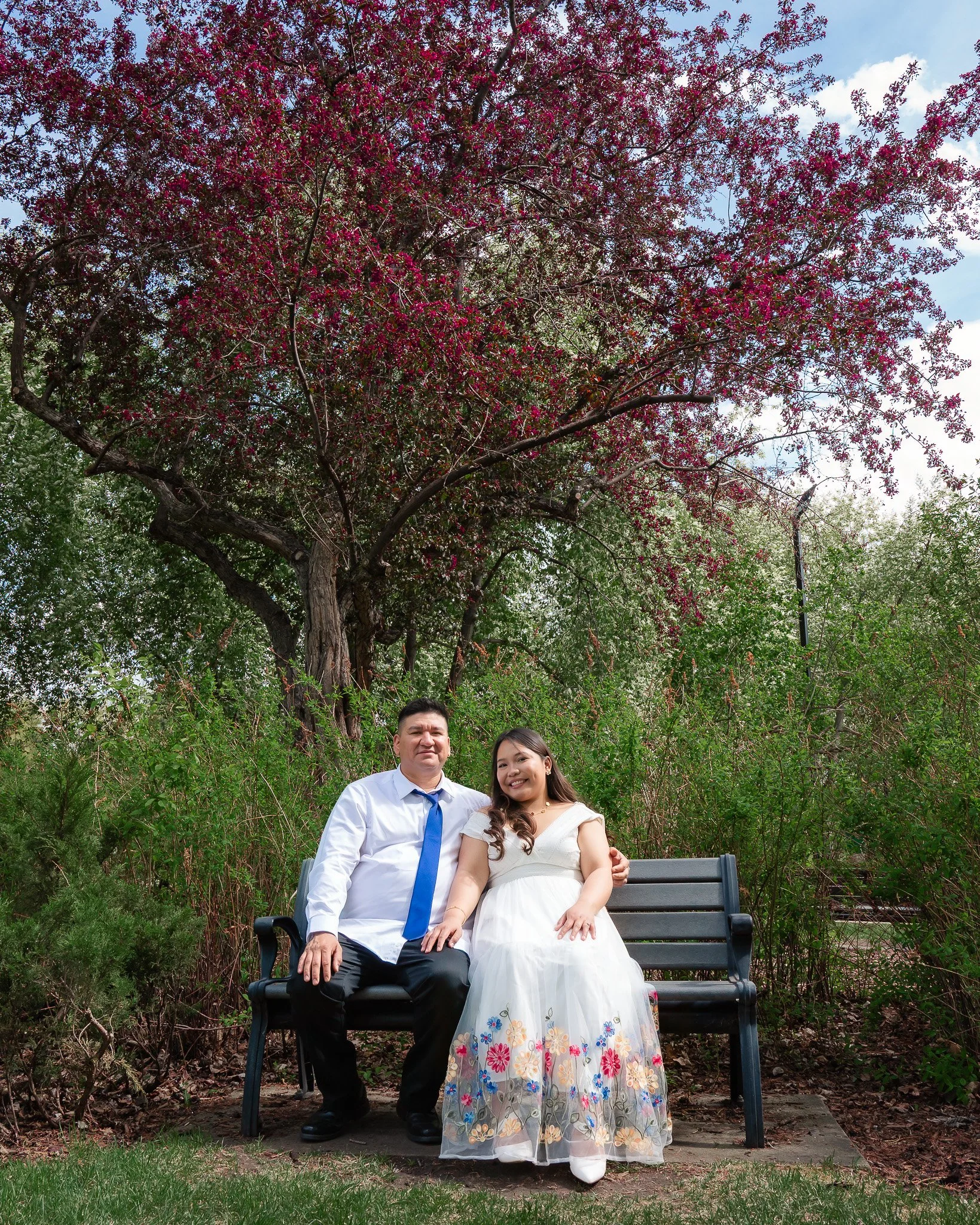 A man and woman sitting on a park bench, dressed formally, with the woman wearing a white dress with colorful floral embroidery and the man in a white shirt and blue tie. They are outdoors with trees and foliage in the background, and a large pink fl