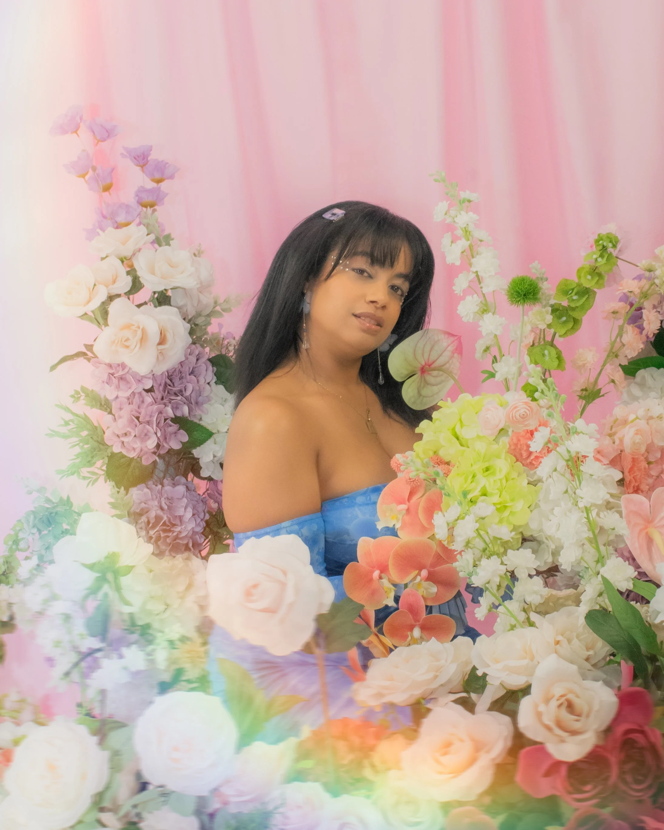 A woman in a blue off-shoulder dress surrounded by pink, purple, white, and green flowers against a pink backdrop.