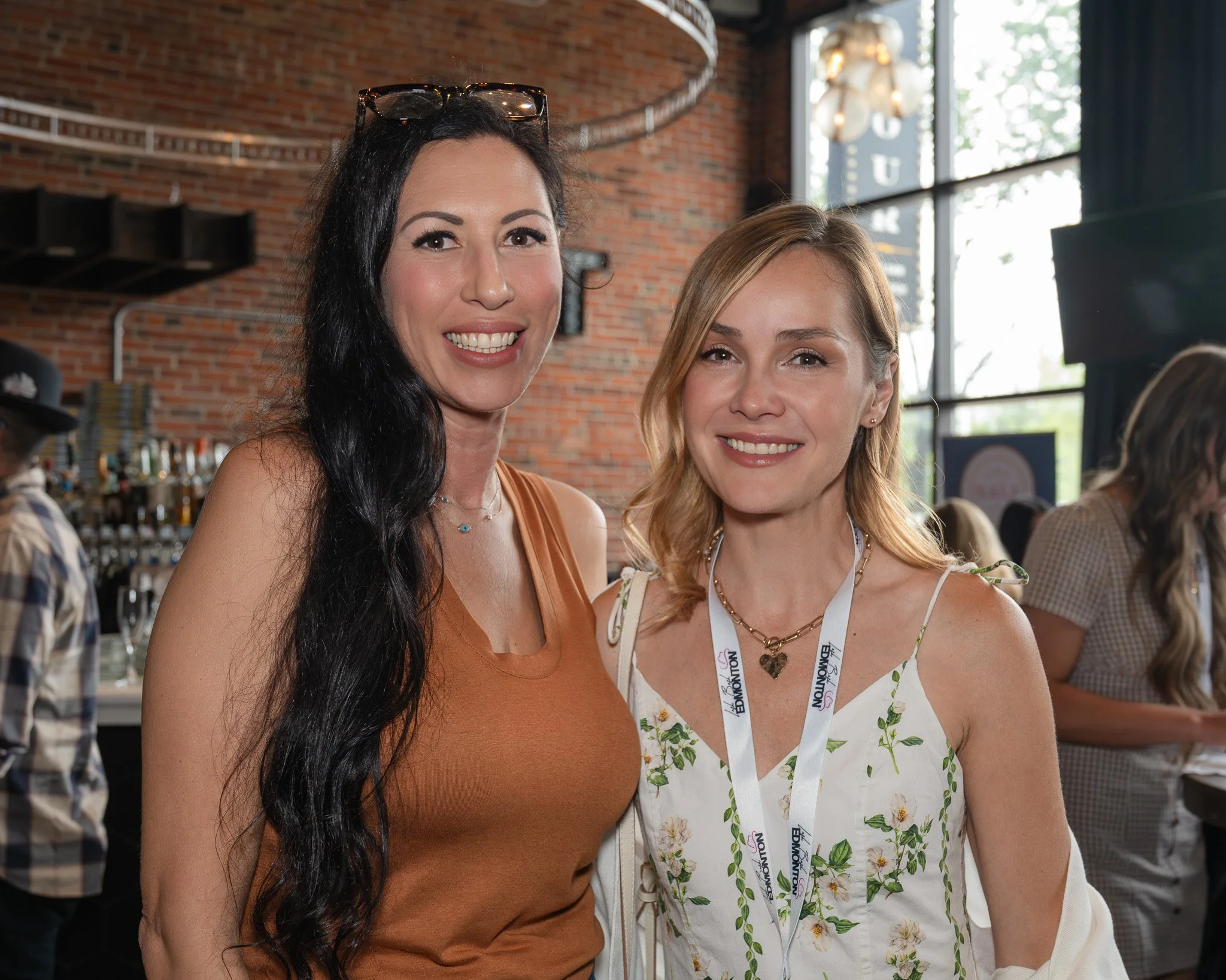Two women smiling at an indoor event, both wearing name badges, with a brick wall and large windows in the background.