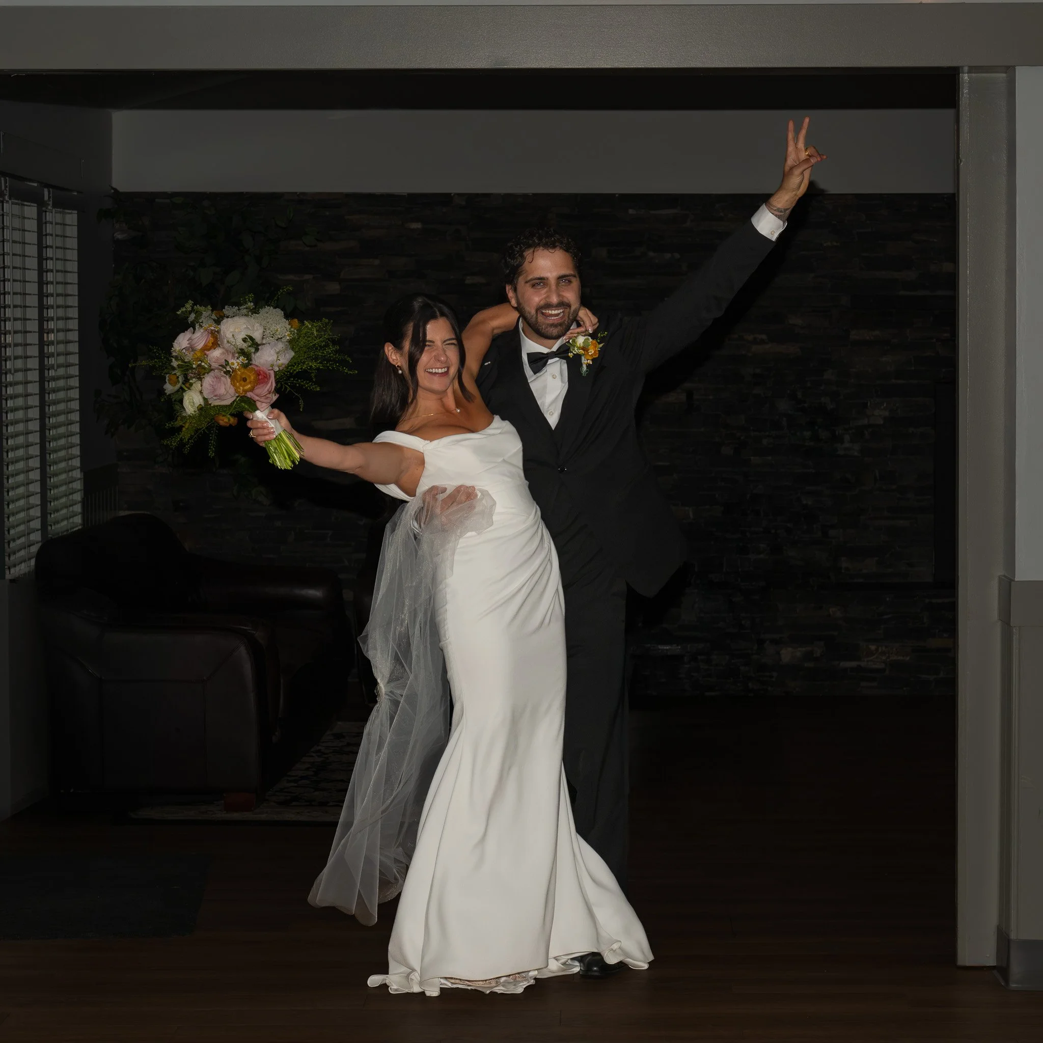 A newlywed couple smiling and posing inside a house. The bride wearing a white wedding dress holding a large bouquet of flowers, the groom in a black tuxedo with a bow tie making a peace sign with his hand.