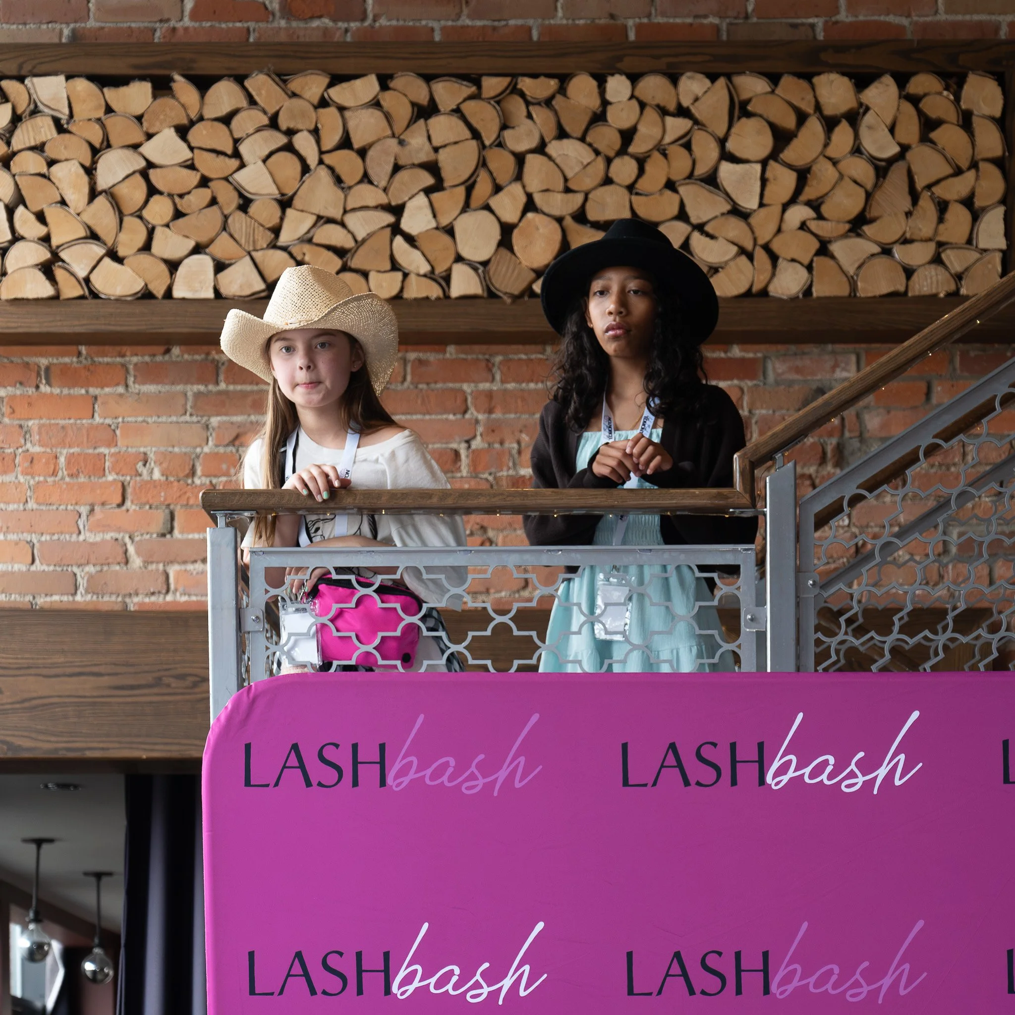 Two young women standing in a clothing store near a display with a pink sign that says "LASH" in black and purple. They are wearing hats, one with a cowboy hat and the other with a black wide-brim hat, with a brick wall and stacked firewood in the ba