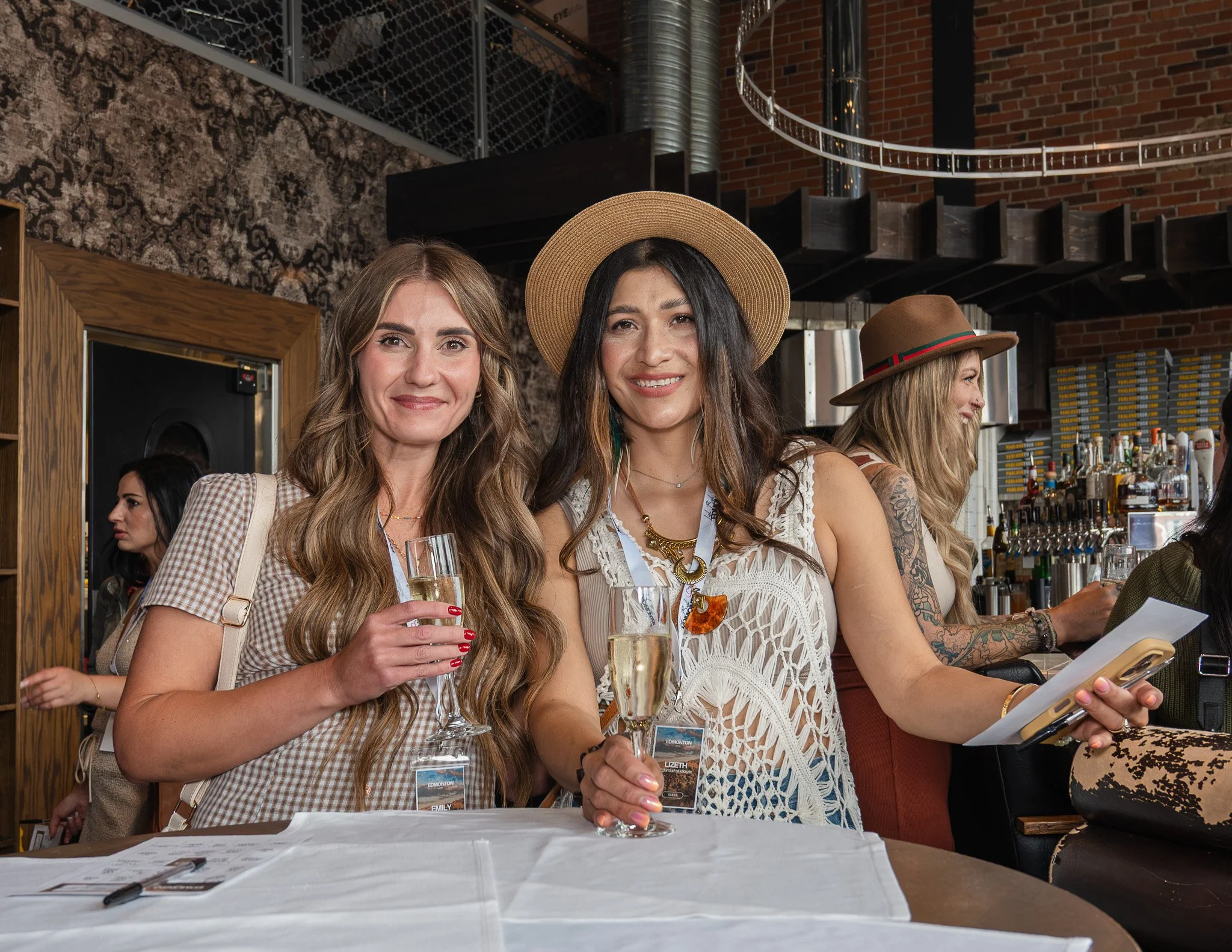 Women attending a social event at a bar, with one woman holding a glass of champagne and smiling at the camera. The women are wearing casual summer clothing and hats, and there is a bar background behind them.