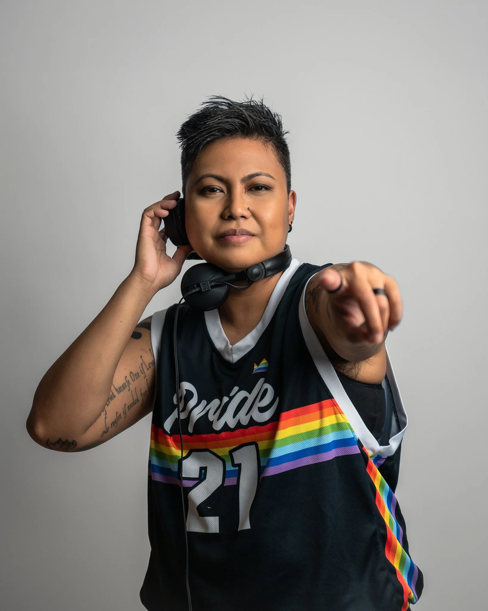 Person with short dark hair wearing a black sleeveless sports jersey with rainbow stripes, holding a headset to the ear, pointing outward, against a plain gray background.