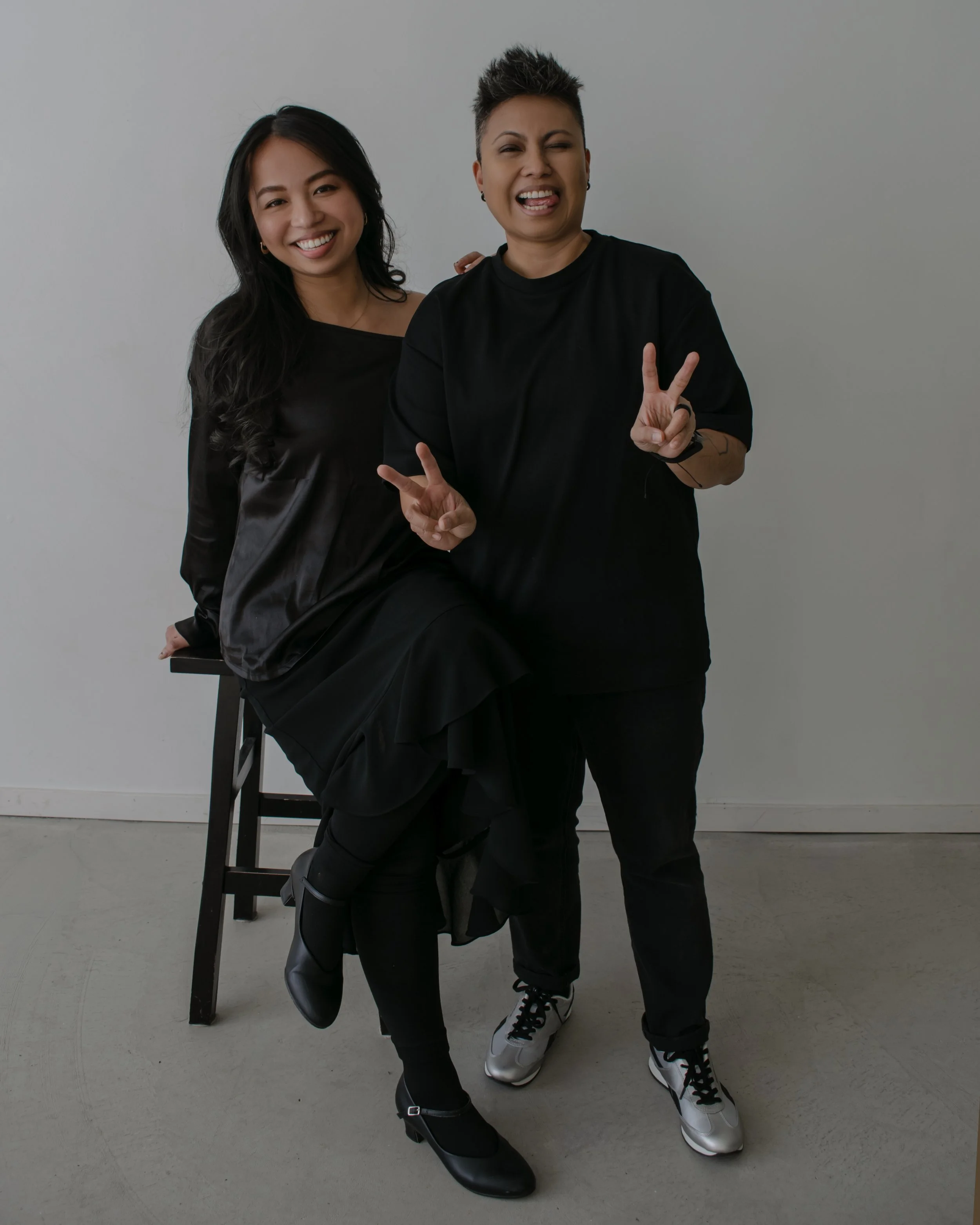 Two women posing indoors against a plain background, one sitting on a chair and the other standing, both smiling and making peace signs.