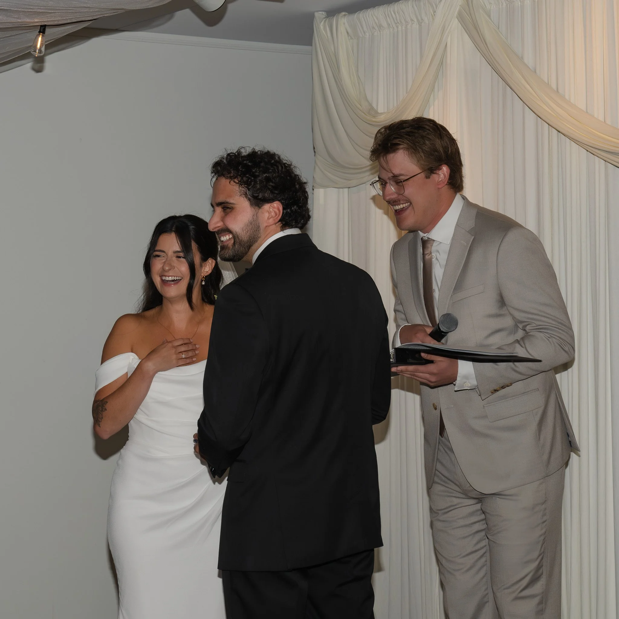 Smiling group of people at a wedding or celebration, standing in front of white curtains and draped fabric, with one person holding a microphone and a clipboard.