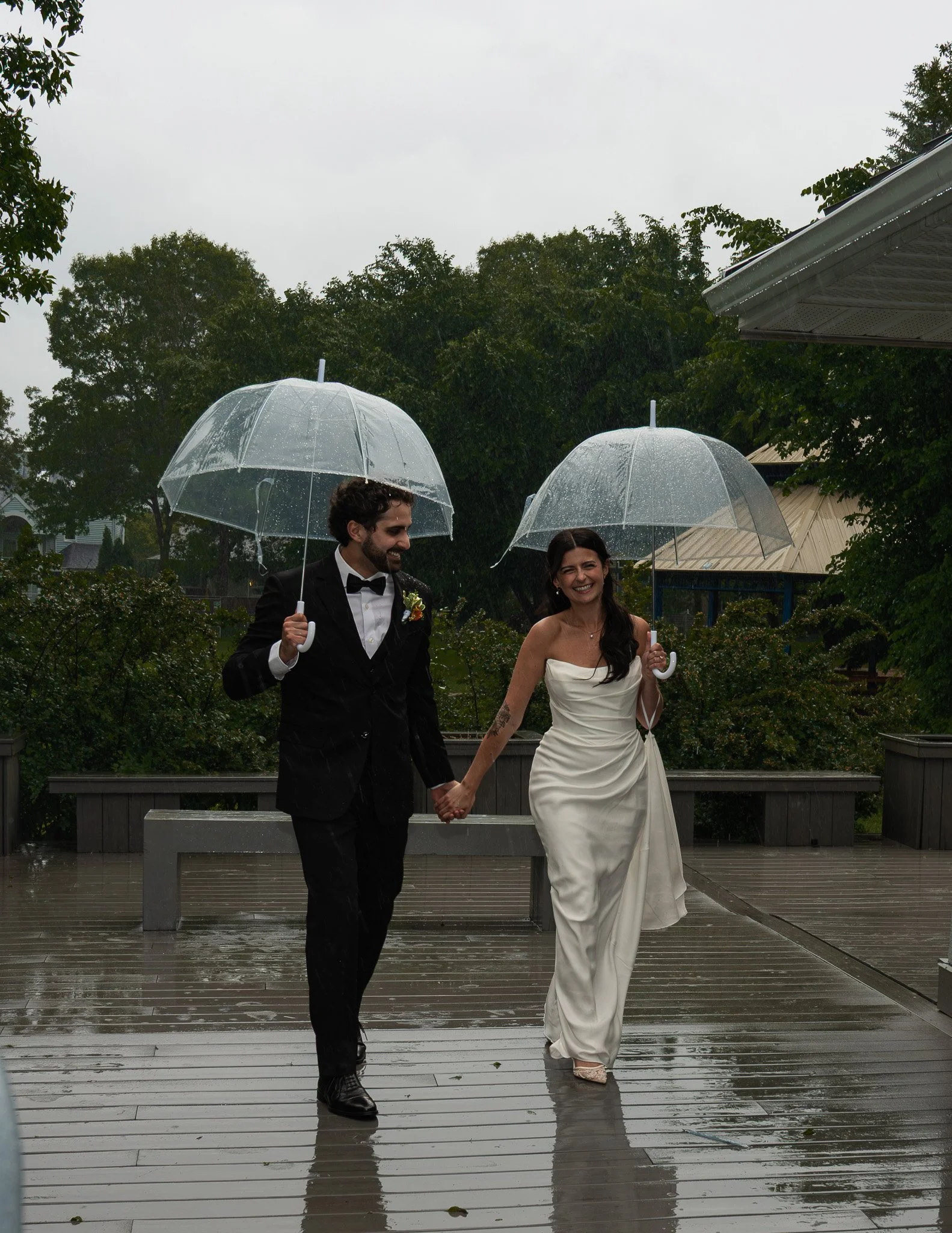 A man and woman dressed in formal attire walking outdoors in the rain, holding clear umbrellas. The man wears a black tuxedo with a bow tie, and the woman wears a strapless white dress. They are smiling and holding hands. The background shows trees, 