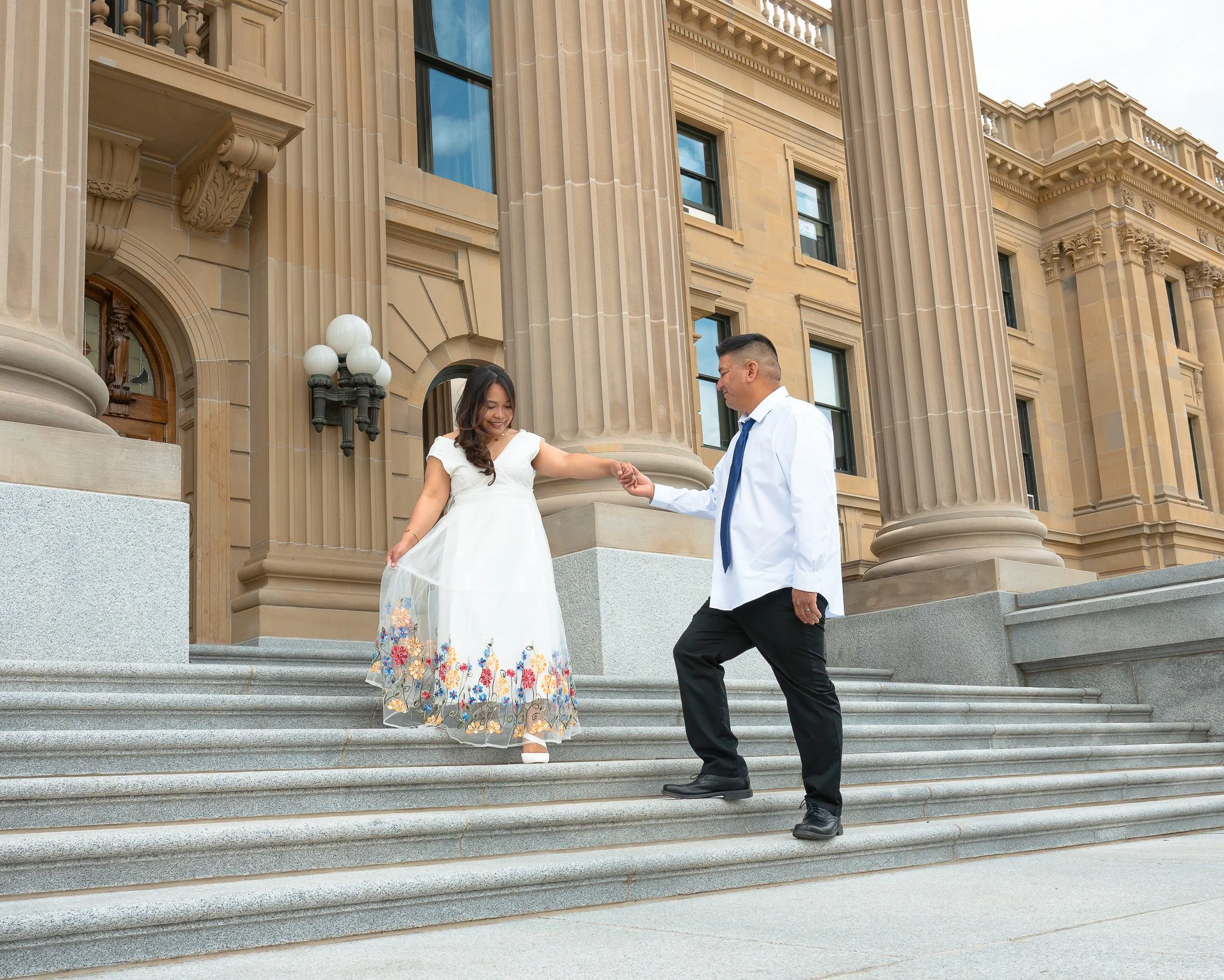 A couple dressed in formal attire standing on the steps of a historic building, holding hands during a wedding ceremony, with a couple of large columns and ornate architecture in the background.
