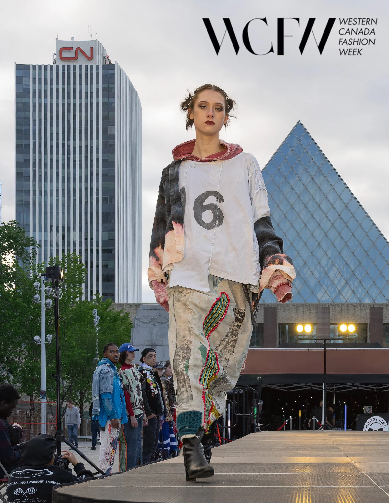 A model walks on an outdoor runway during Western Canada Fashion Week. She wears a distressed, oversized shirt with the number 6 printed on it, and colorful, patterned embroidery and knit details on her pants. The background features tall modern buil