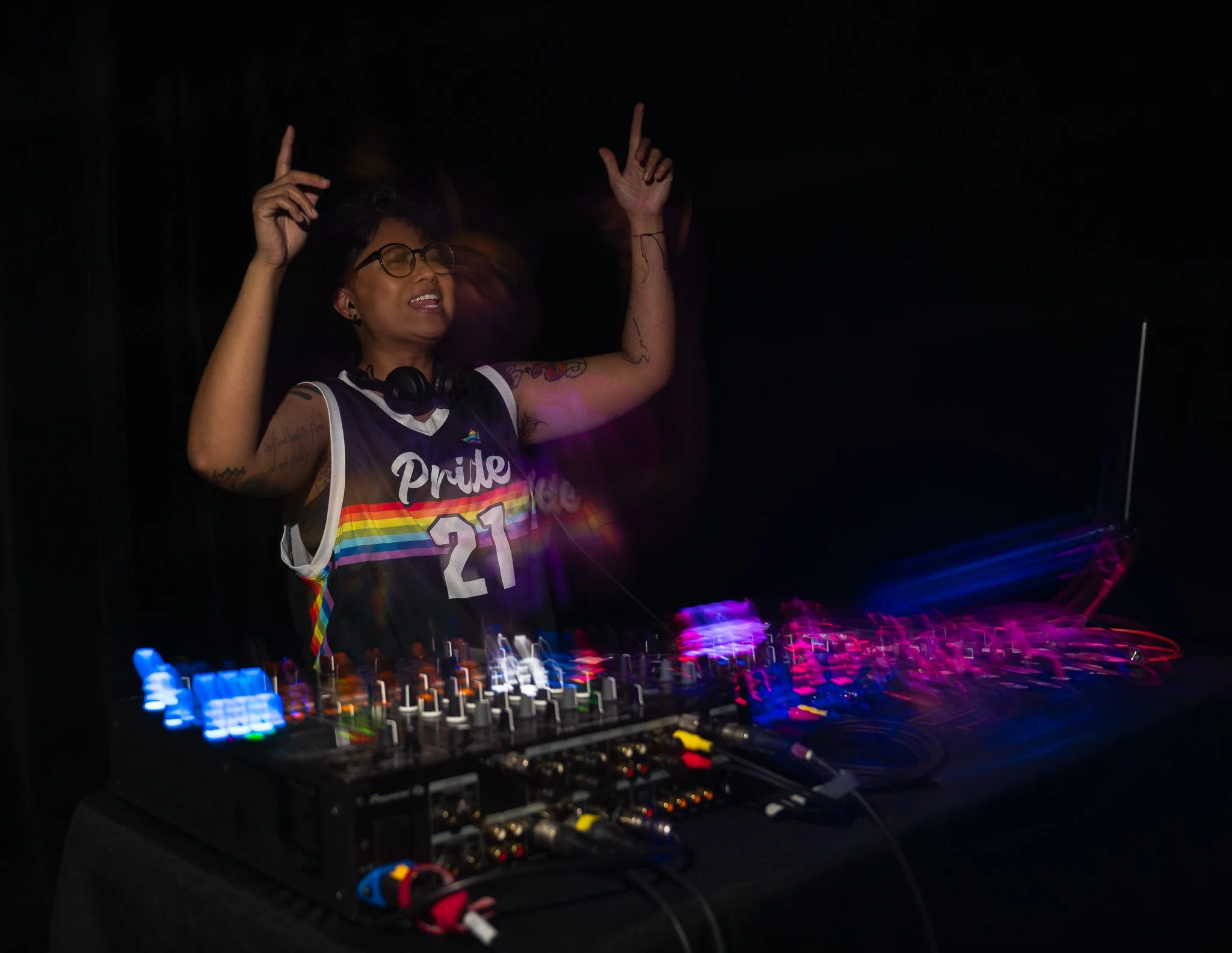 A woman with tattoos and glasses, wearing a rainbow pride tank top, is DJing at a dark party with colorful lights reflecting off the equipment.