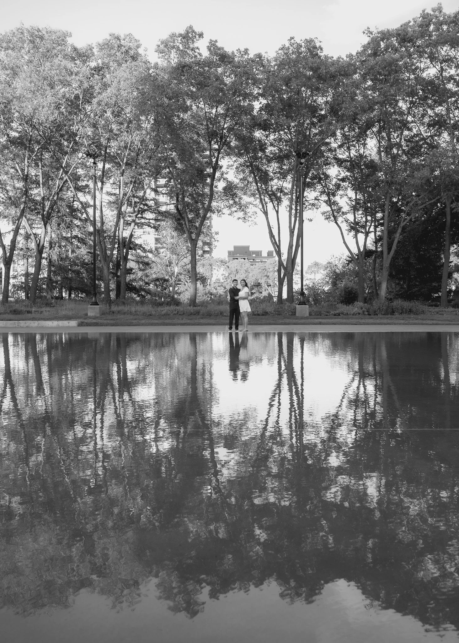 A black-and-white photo of a couple standing near a pond, with a line of tall trees in the background and buildings visible behind the trees.