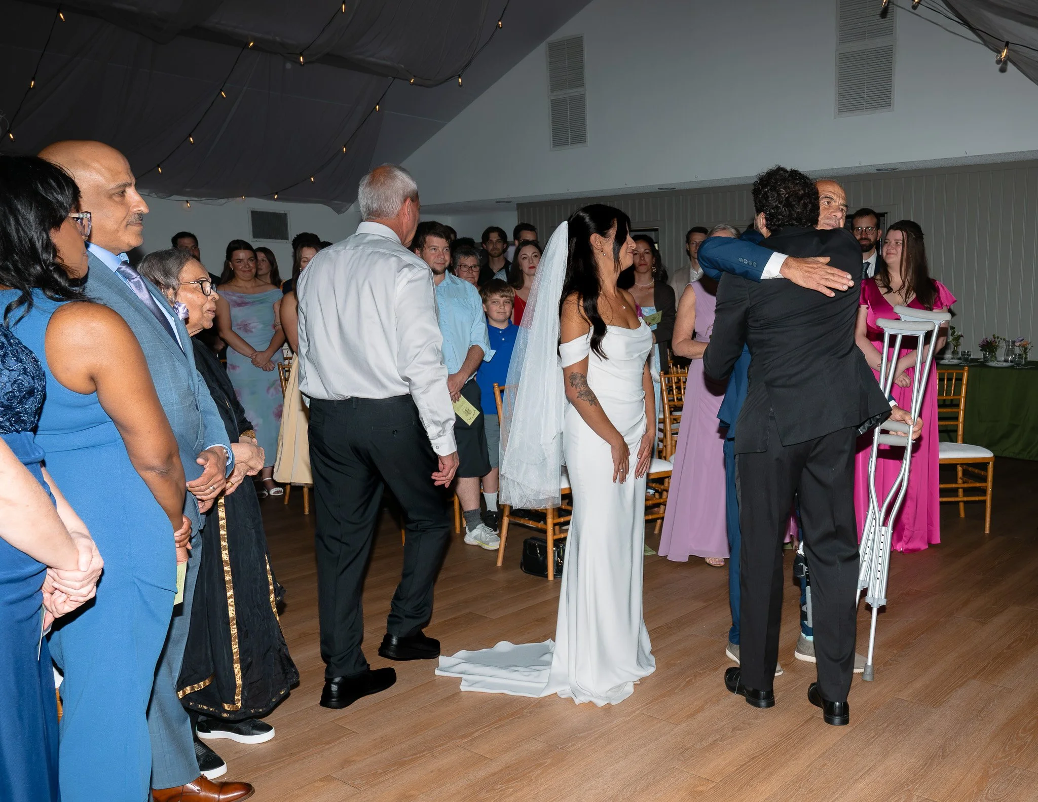 A group of people, including a woman in a white wedding gown and a man in a black suit, are standing closely together at a wedding reception. The seating arrangement and table decorations suggest a formal indoor celebration.