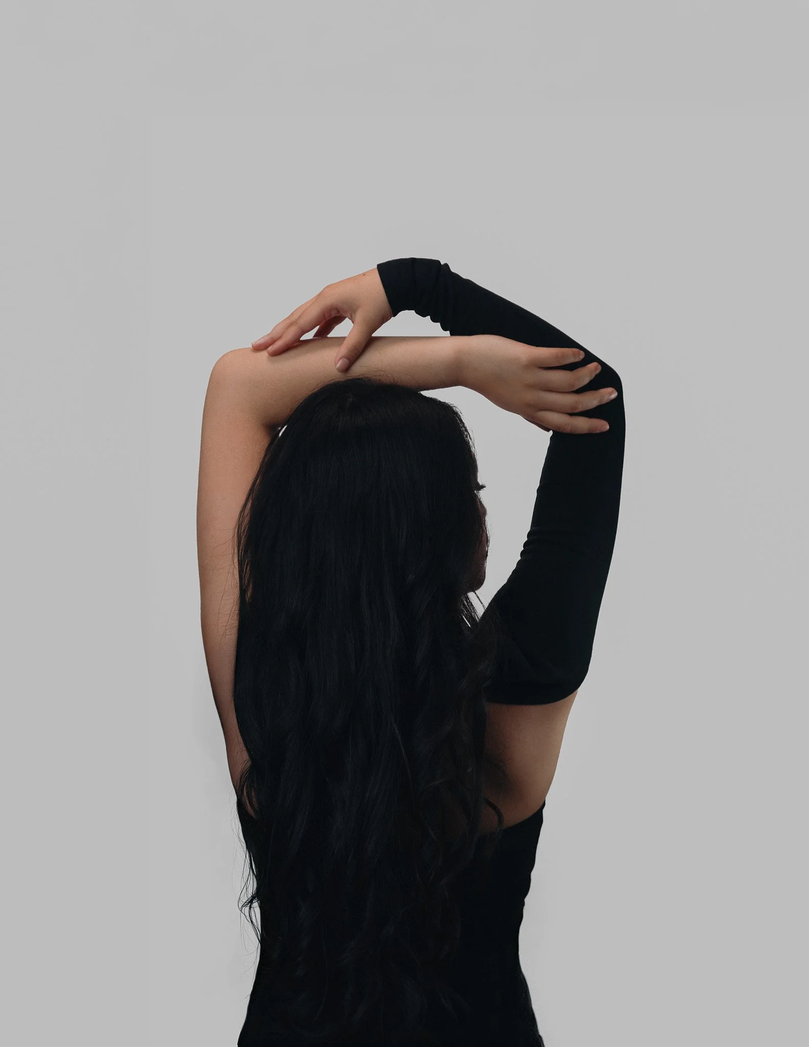 A woman with long black hair is stretching her arms over her head, with one arm bent and the other straight, against a plain gray background. She is wearing a black long-sleeve shirt.